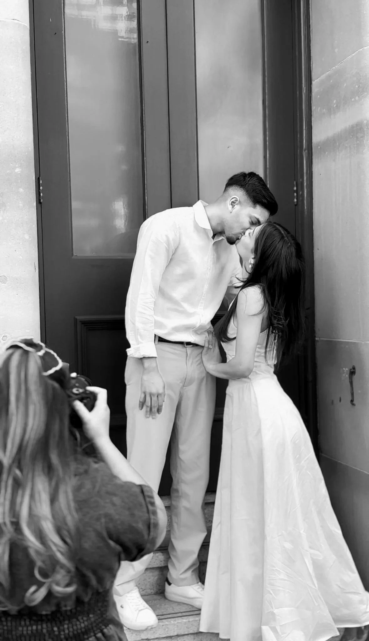 A black-and-white photo of a couple kissing in front of a door while another person takes a picture of them.