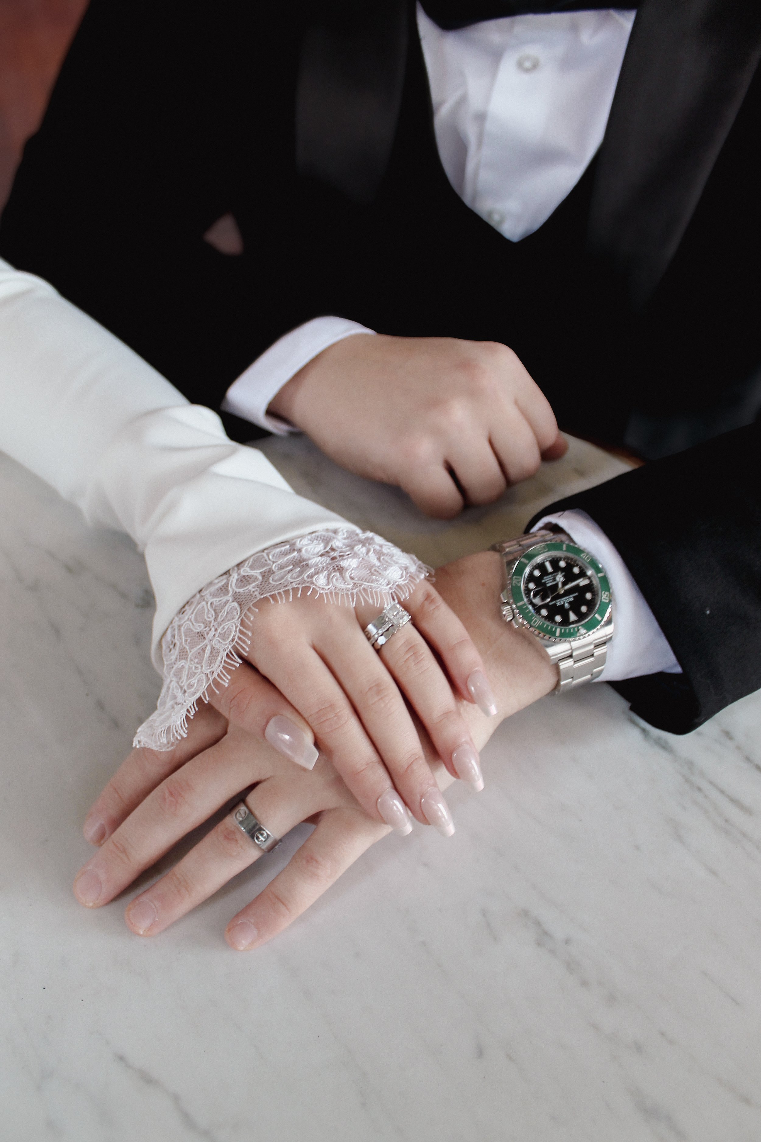 Close-up of a couple's hands, showing wedding rings, resting on a marble surface. The woman wears a white lace sleeve and a diamond engagement ring, the man wears a black tuxedo with a wristwatch.