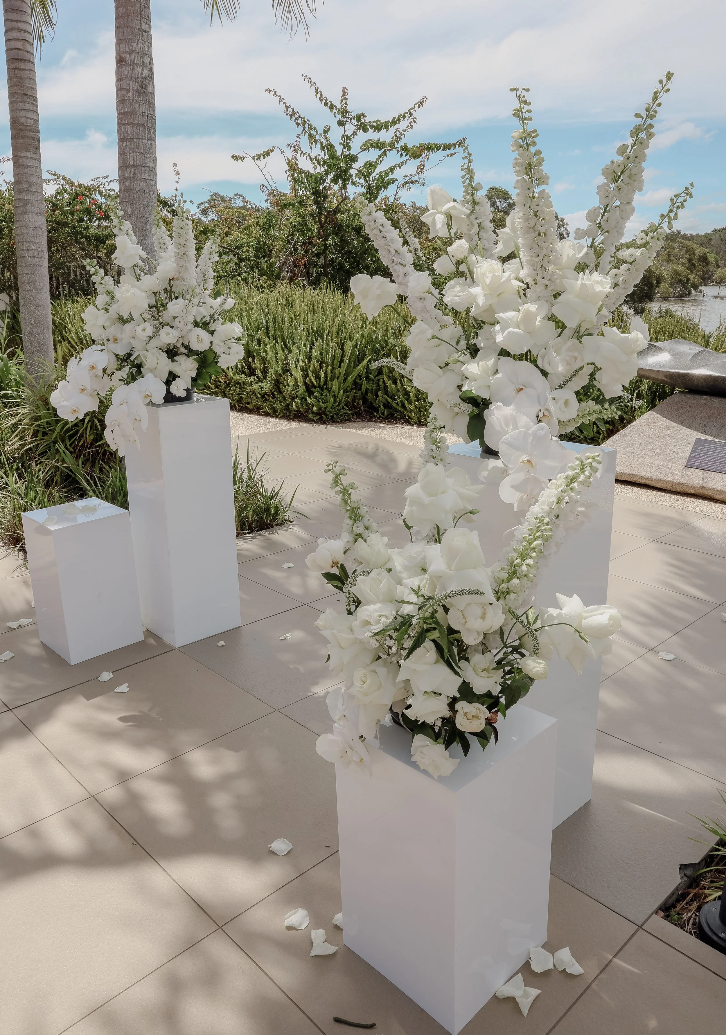 White flower arrangements on white pedestals at an outdoor event.