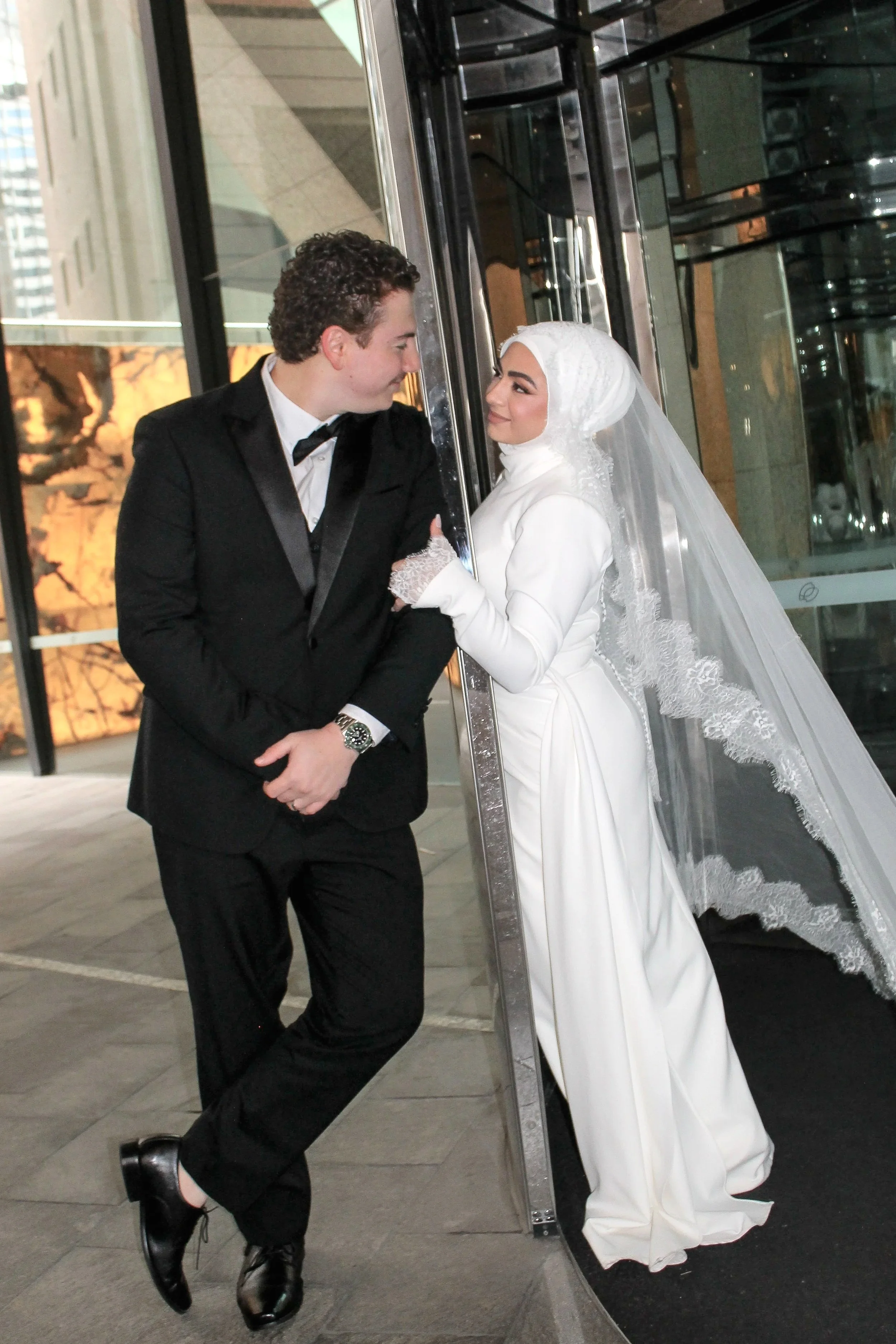 Bride and groom in wedding attire sharing a moment outside a modern building with large glass windows.