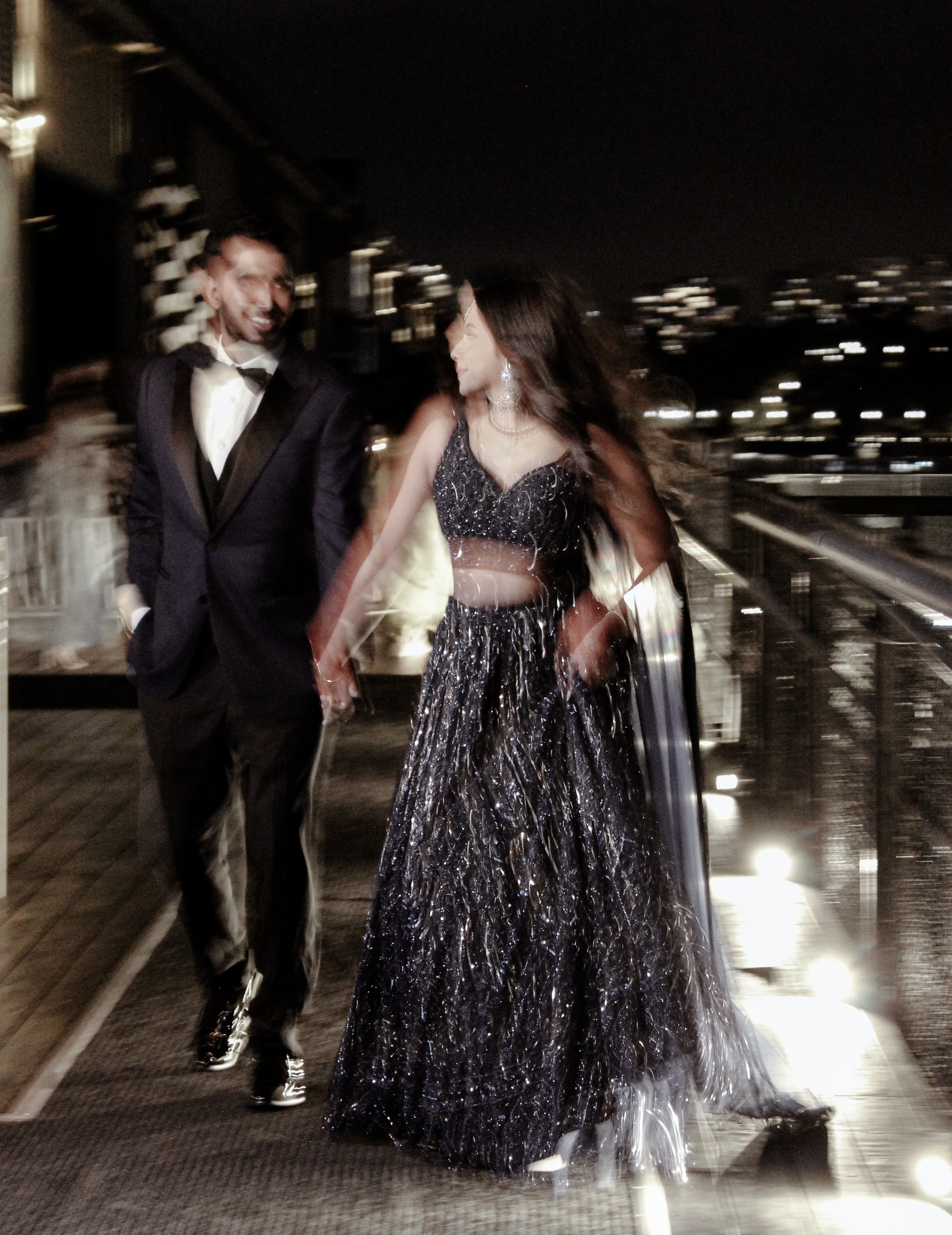 A man and woman dressed in formal evening attire walking on an outdoor balcony at night, with city lights in the background.