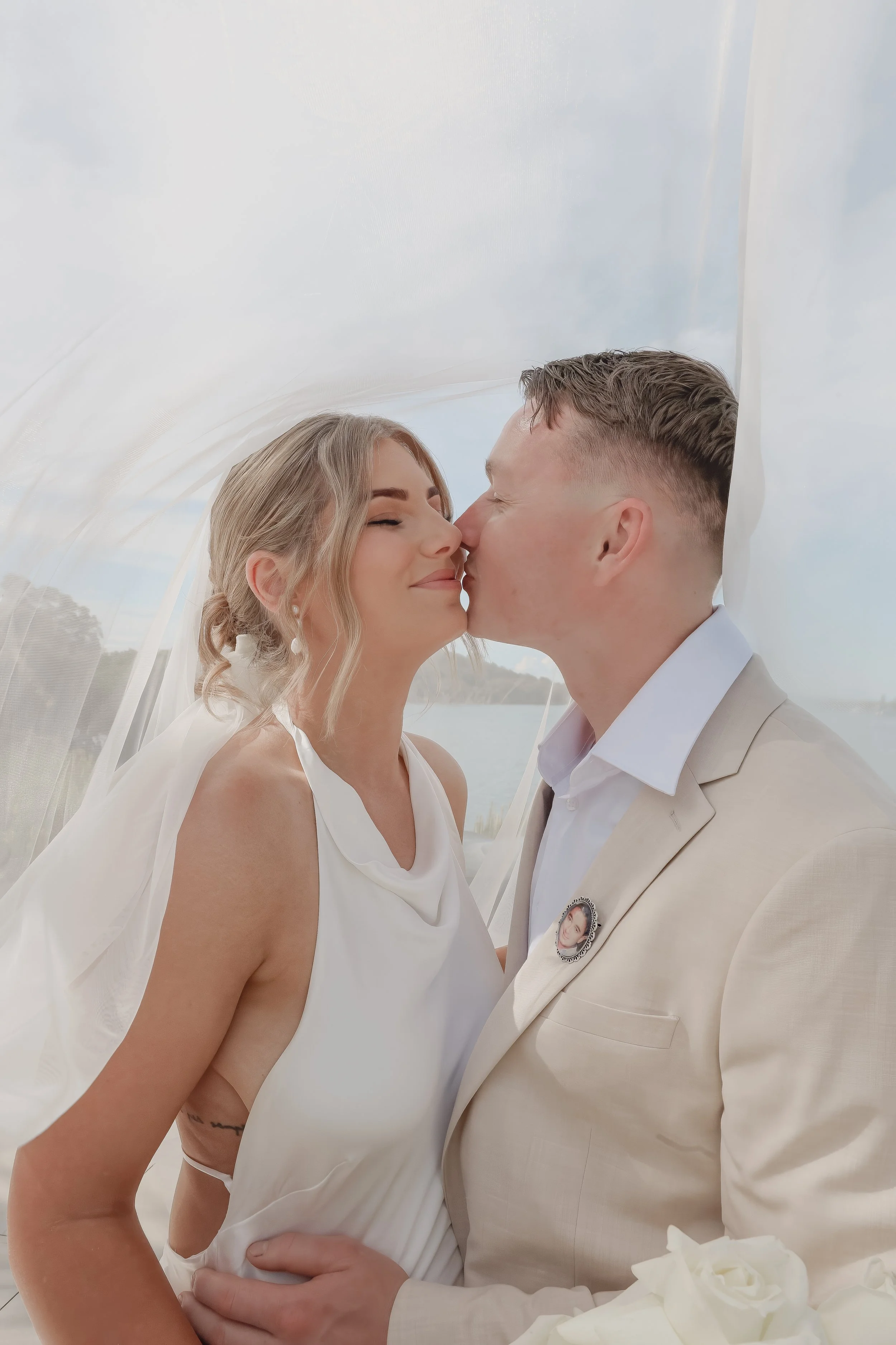 A bride and groom sharing a kiss at their wedding, with the bride smiling and the groom gently holding her waist, in a scenic outdoor setting near water.