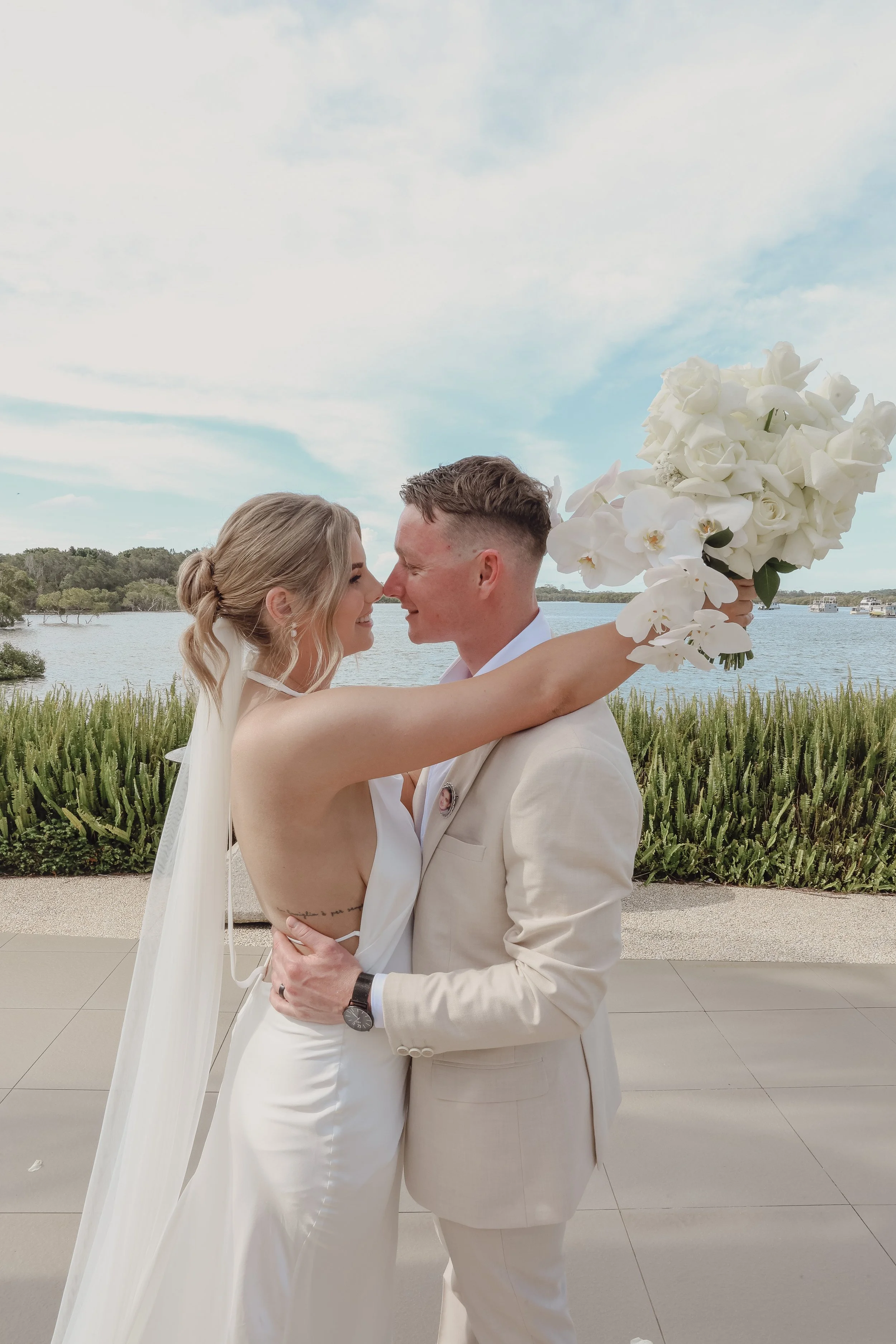 A newlywed couple sharing an intimate moment outdoors with a lake and sky in the background. The bride is holding a large bouquet of white flowers, and they are smiling with foreheads touching.