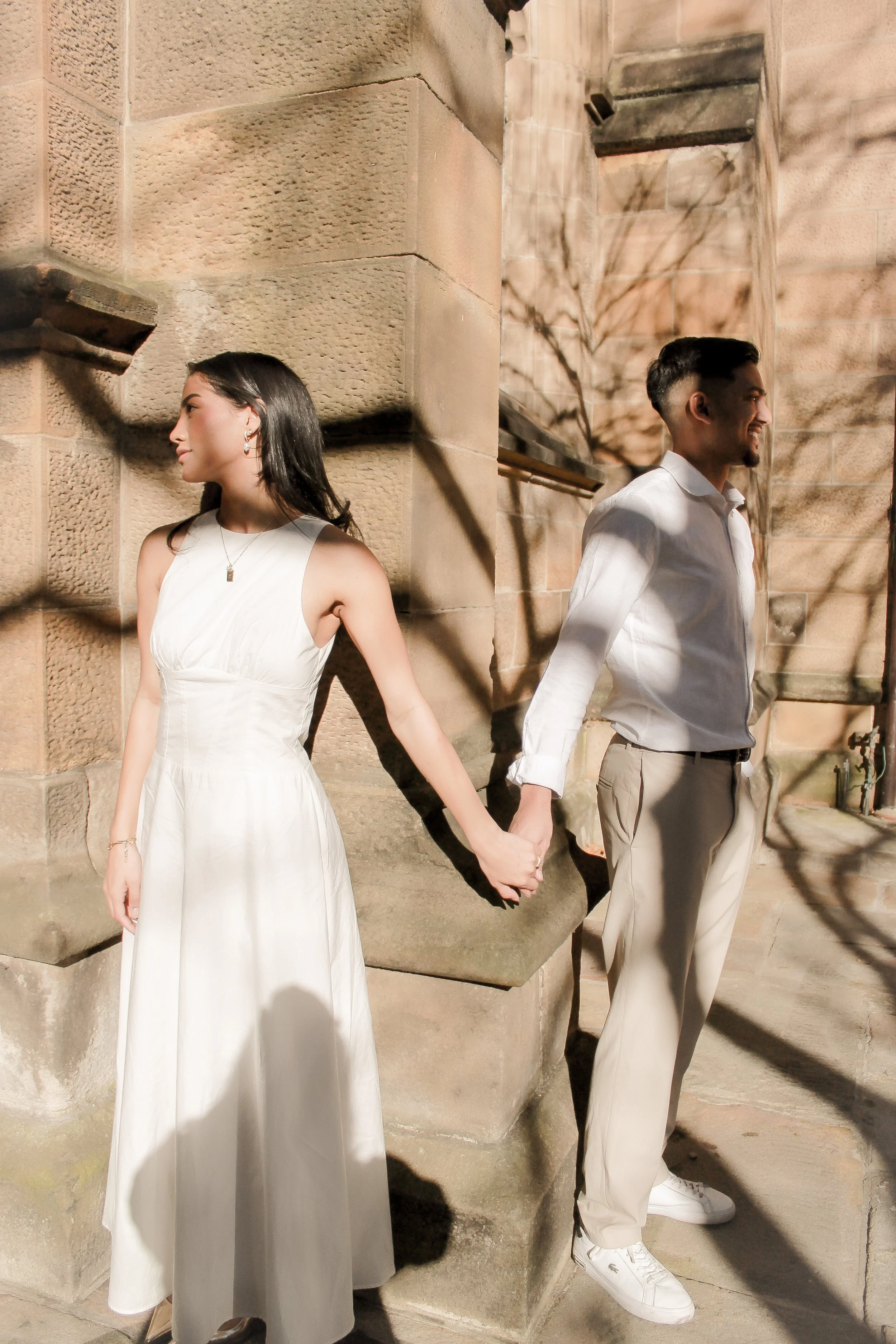 A young woman and a young man hold hands and stand back-to-back, outdoors in sunlight near a stone building, both smiling.