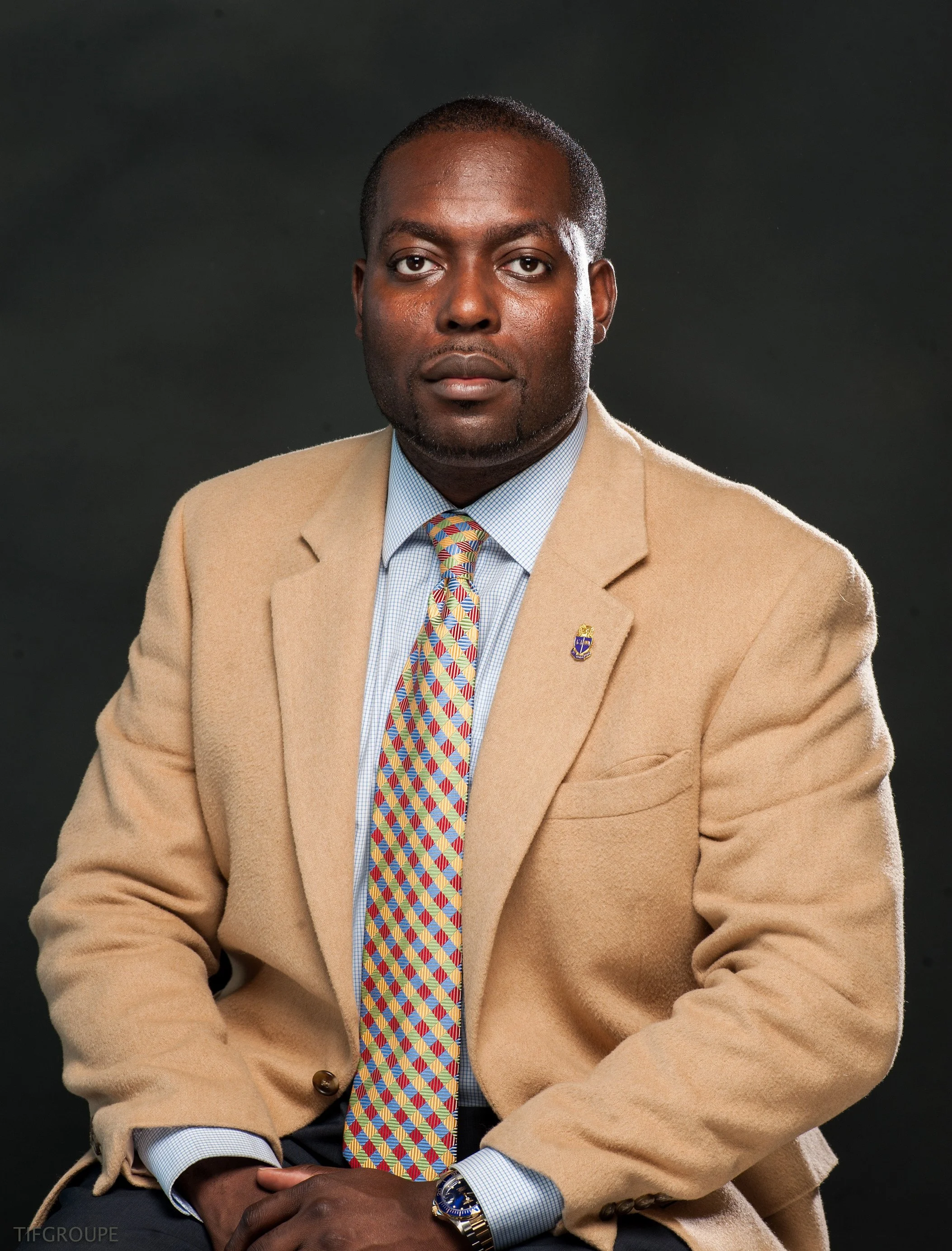 Portrait of a man wearing a beige blazer, a blue shirt, and a colorful geometric tie against a dark background.
