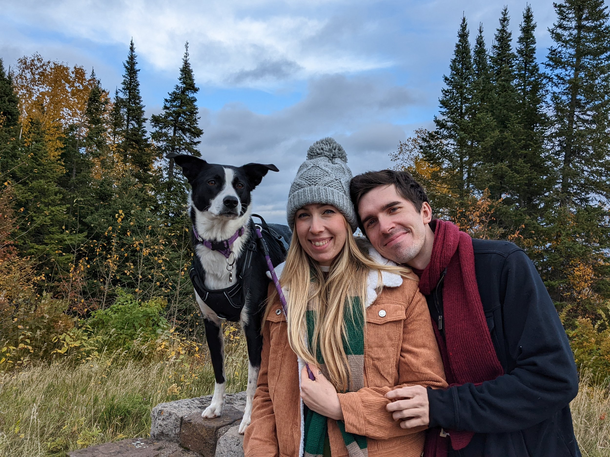 A couple and their dog outdoors in a forested area during autumn. The woman is wearing a gray beanie and a brown jacket with a green scarf, and the man is wearing a dark jacket with a maroon scarf. The dog, a black and white breed, is standing next to them on a rock.
