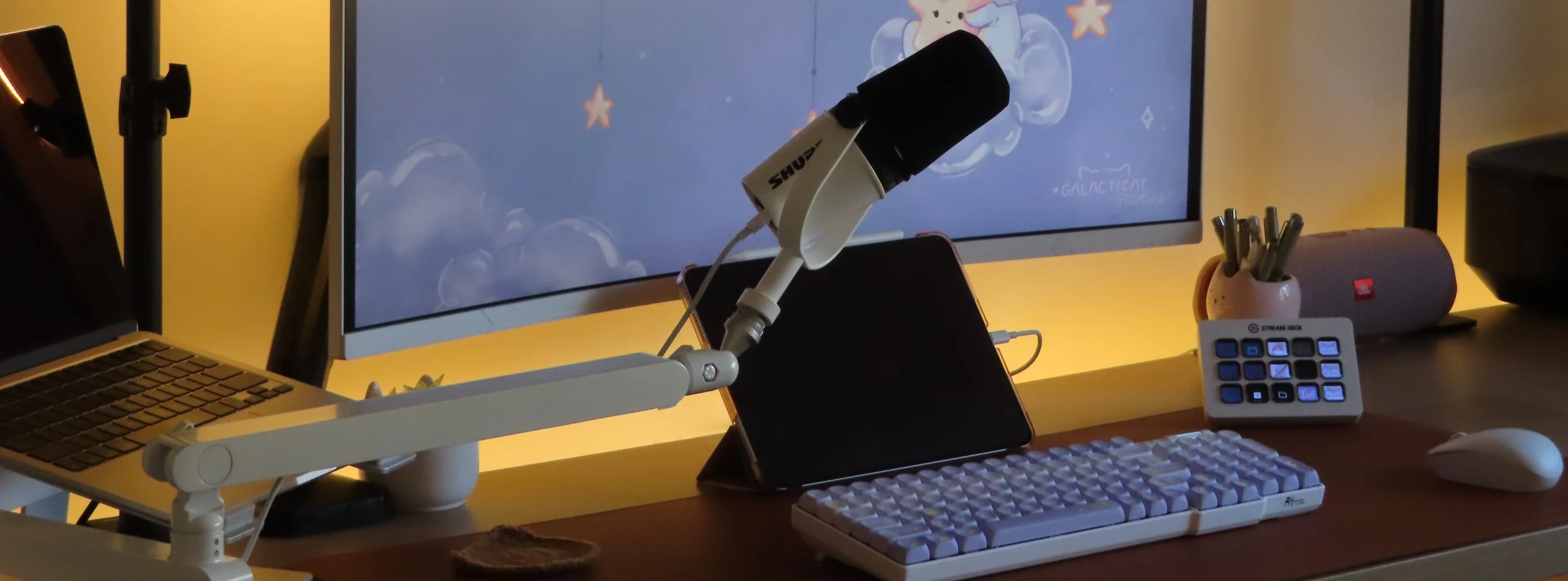 A computer desk setup featuring a monitor with a cartoon-themed background, an adjustable arm holding a microphone, a laptop, a tablet, a mechanical keyboard, a mouse, a small stream deck, a pen holder with pens, a speaker, and a partially visible laptop.