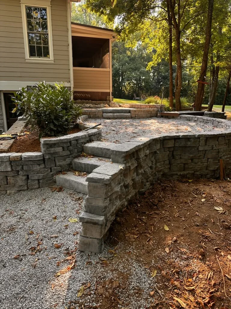 Stone retaining wall and steps under construction near house with beige siding and surrounded by trees.