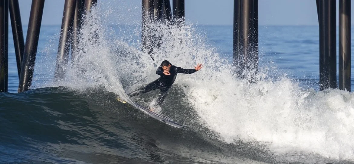 Surfer in a wetsuit riding a wave near a pier.