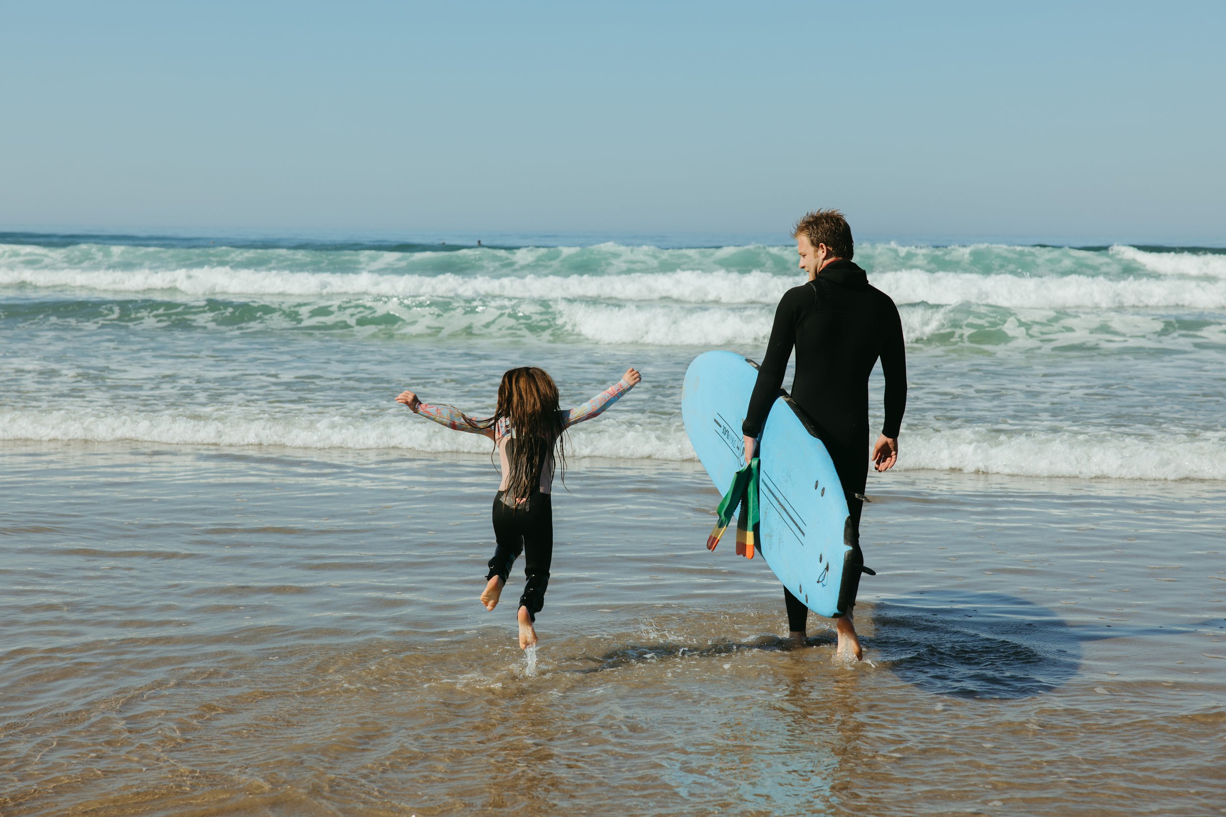 A man and a girl with surfboards on a sandy beach during sunset, with a pier and hills in the background.