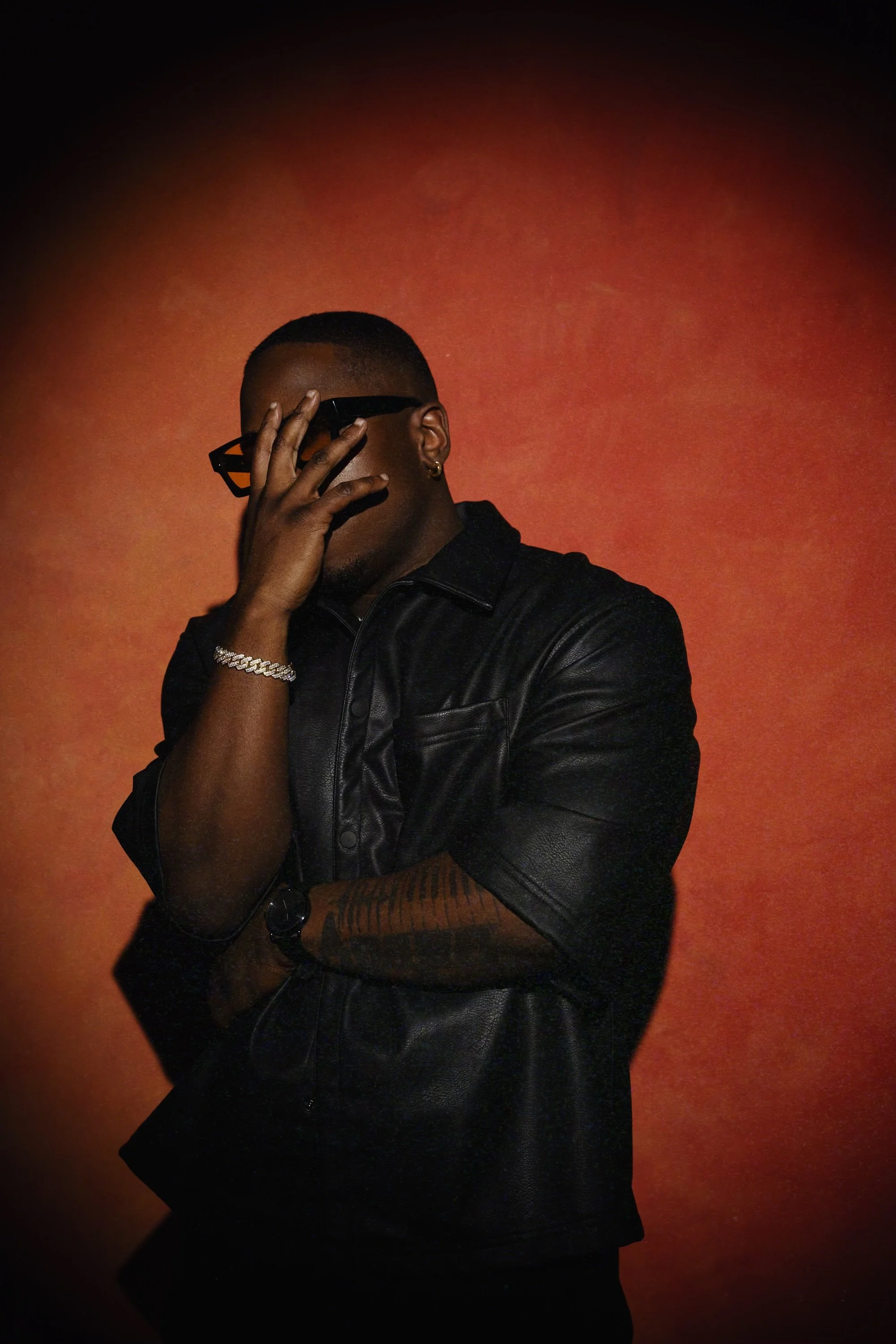 Aaron Hines, contemporary R&B artist, wearing a black leather outfit and sunglasses, standing against a red background in a moody portrait