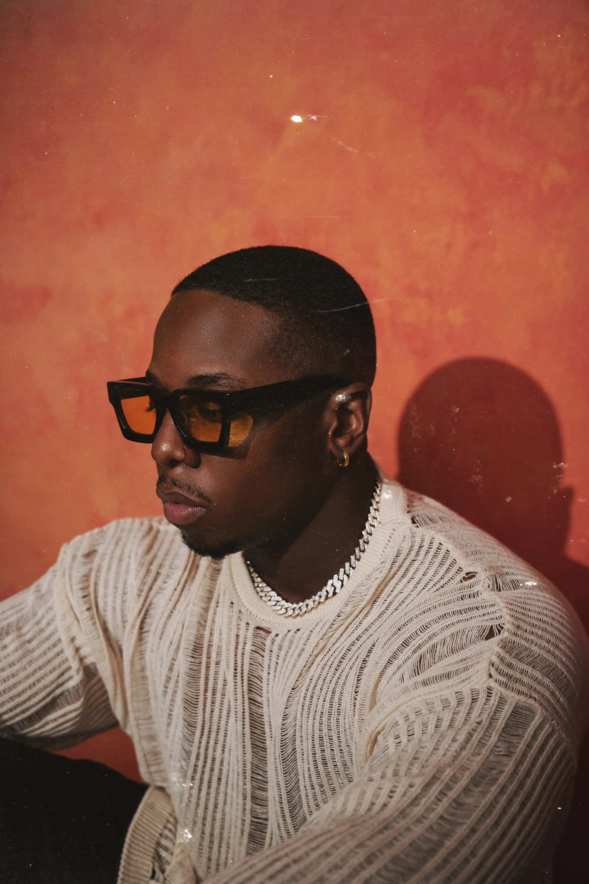 Aaron Hines, R&B and soul artist, wearing a white textured sweater and sunglasses, seated against a warm orange background