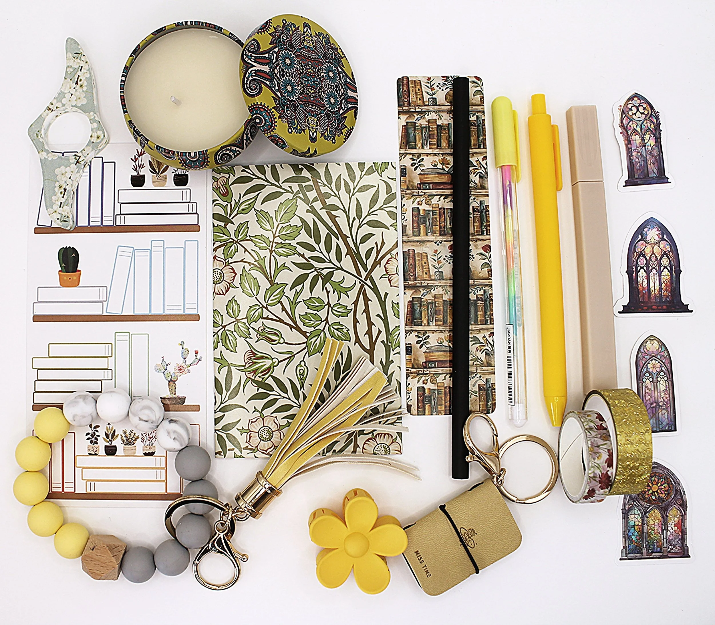 Assorted stationery and decorative items including candles, notebooks, pens, washi tapes, stickers, keychains, a bracelet, and book-themed paper cutouts arranged on a white surface.