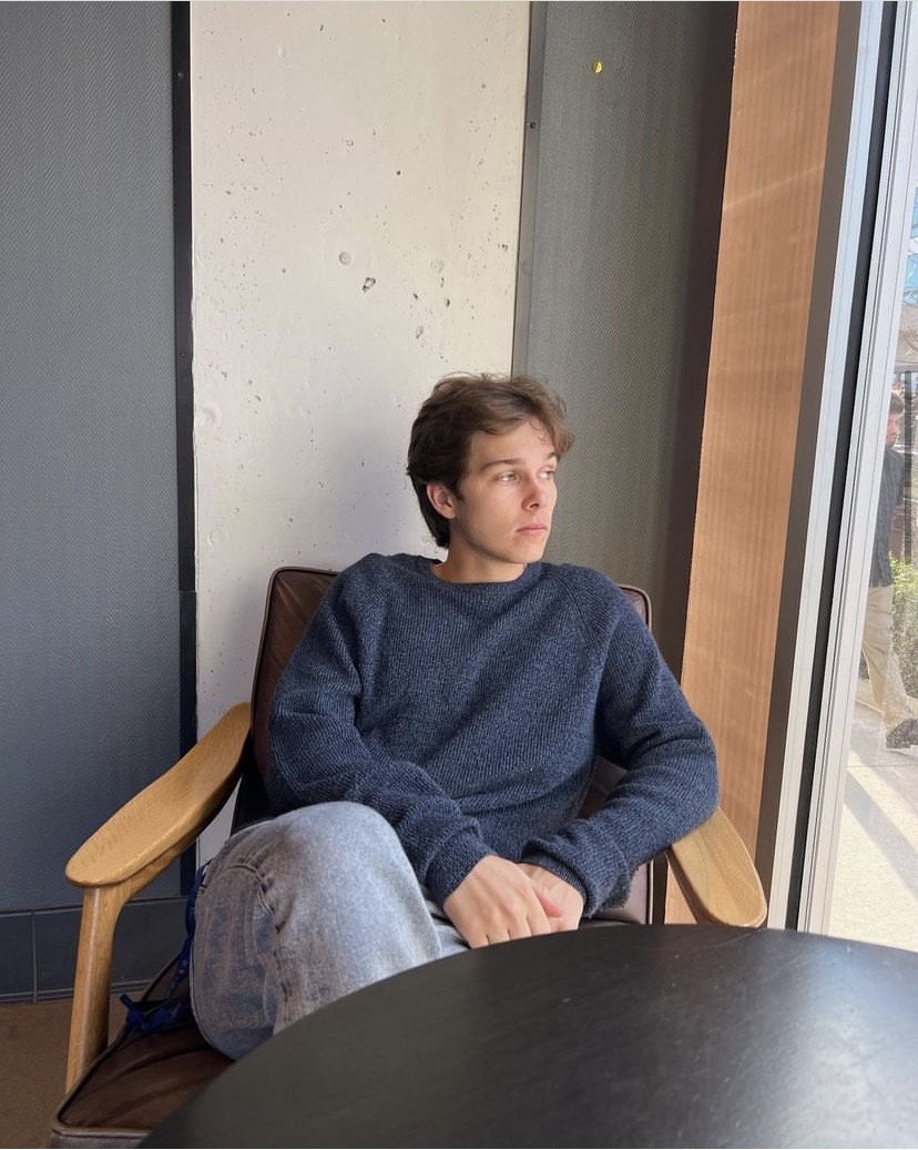 A young person with short brown hair wearing a blue sweater and light-colored jeans sits in a wooden armchair near a large window, looking outside thoughtfully.