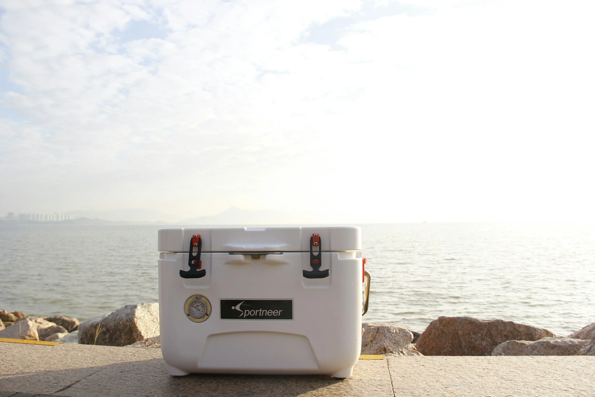 White cooler on a concrete surface by the water, with rocks along the shoreline and an overcast sky in the background.