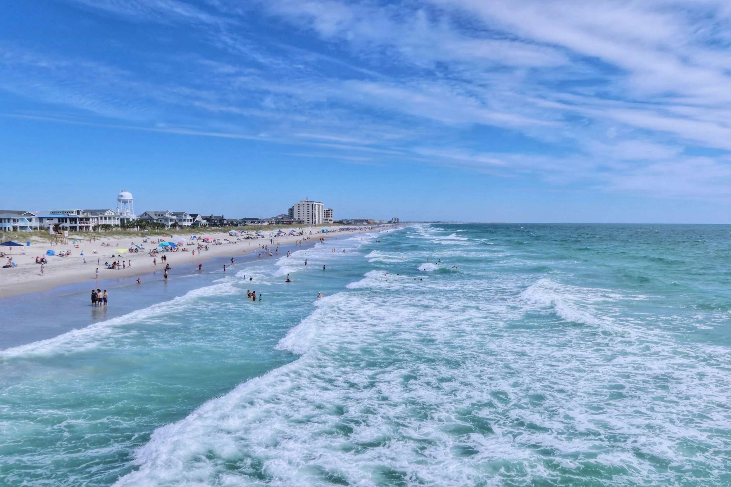 Beach scene with people swimming and relaxing along the shoreline, with residential buildings and a water tower in the background, under a partly cloudy blue sky.