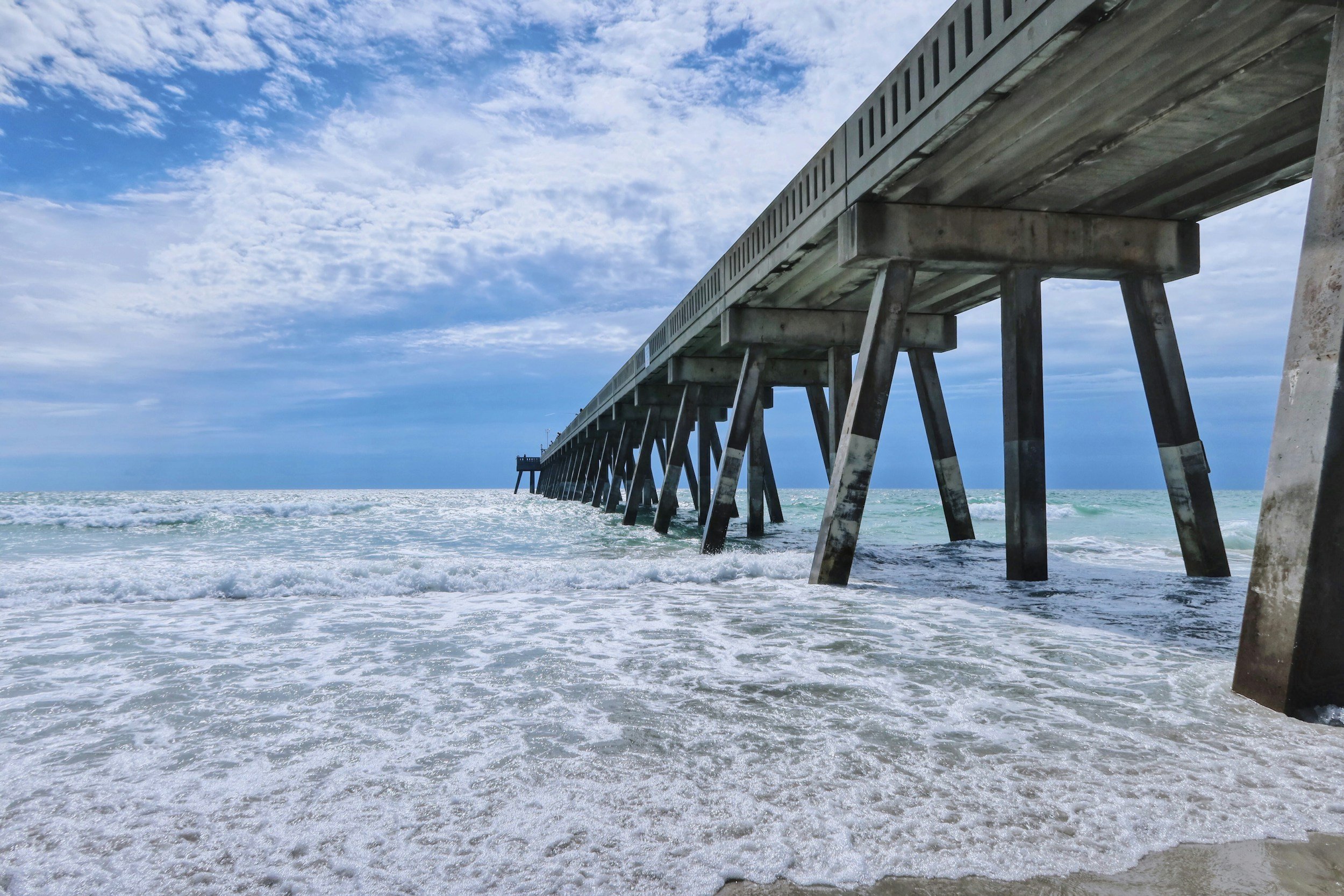 View of a concrete pier extending into the ocean, with waves crashing against the shore and a partly cloudy sky overhead.