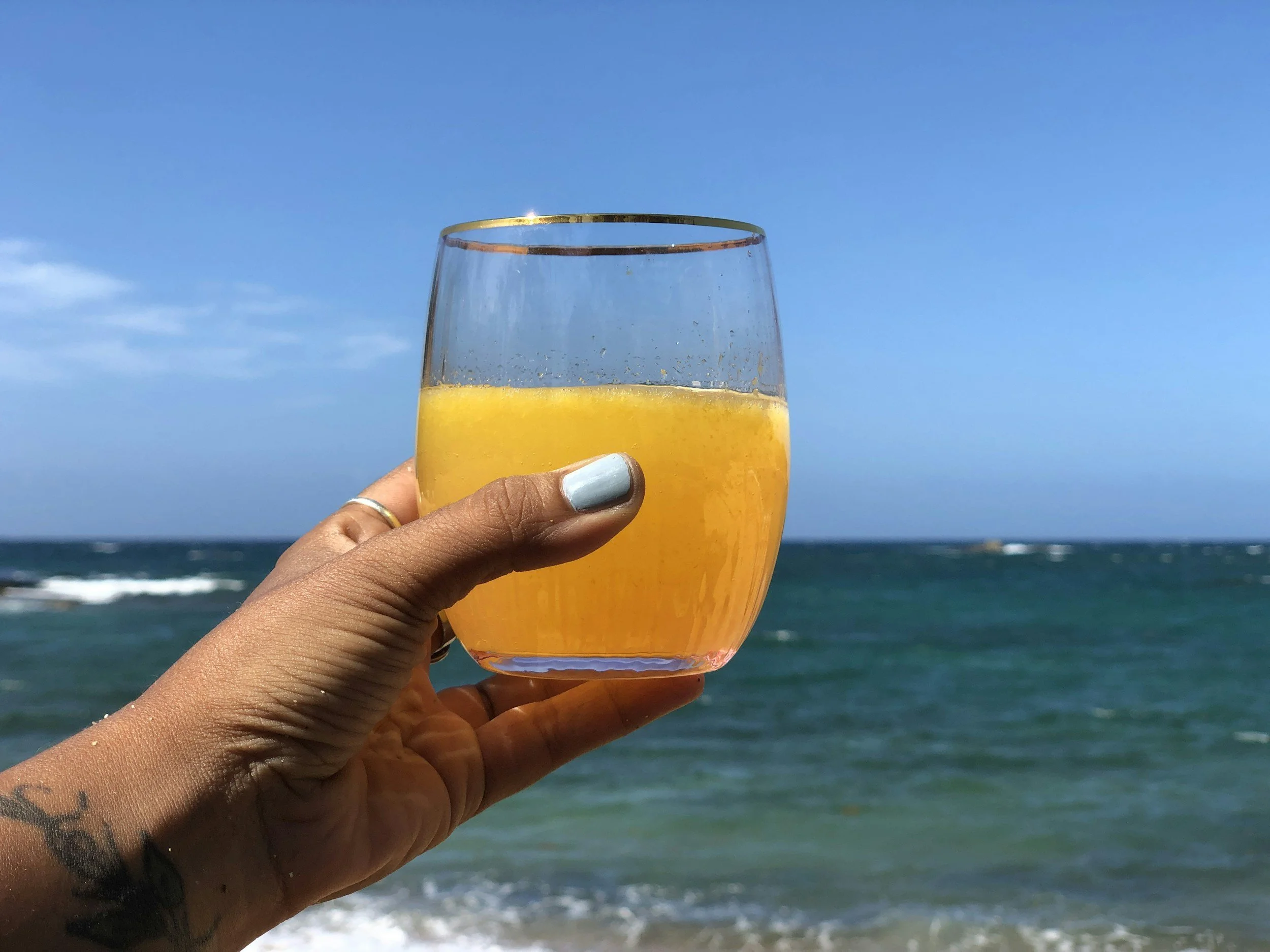 Hand holding a glass of orange juice against a backdrop of the ocean and a blue sky.