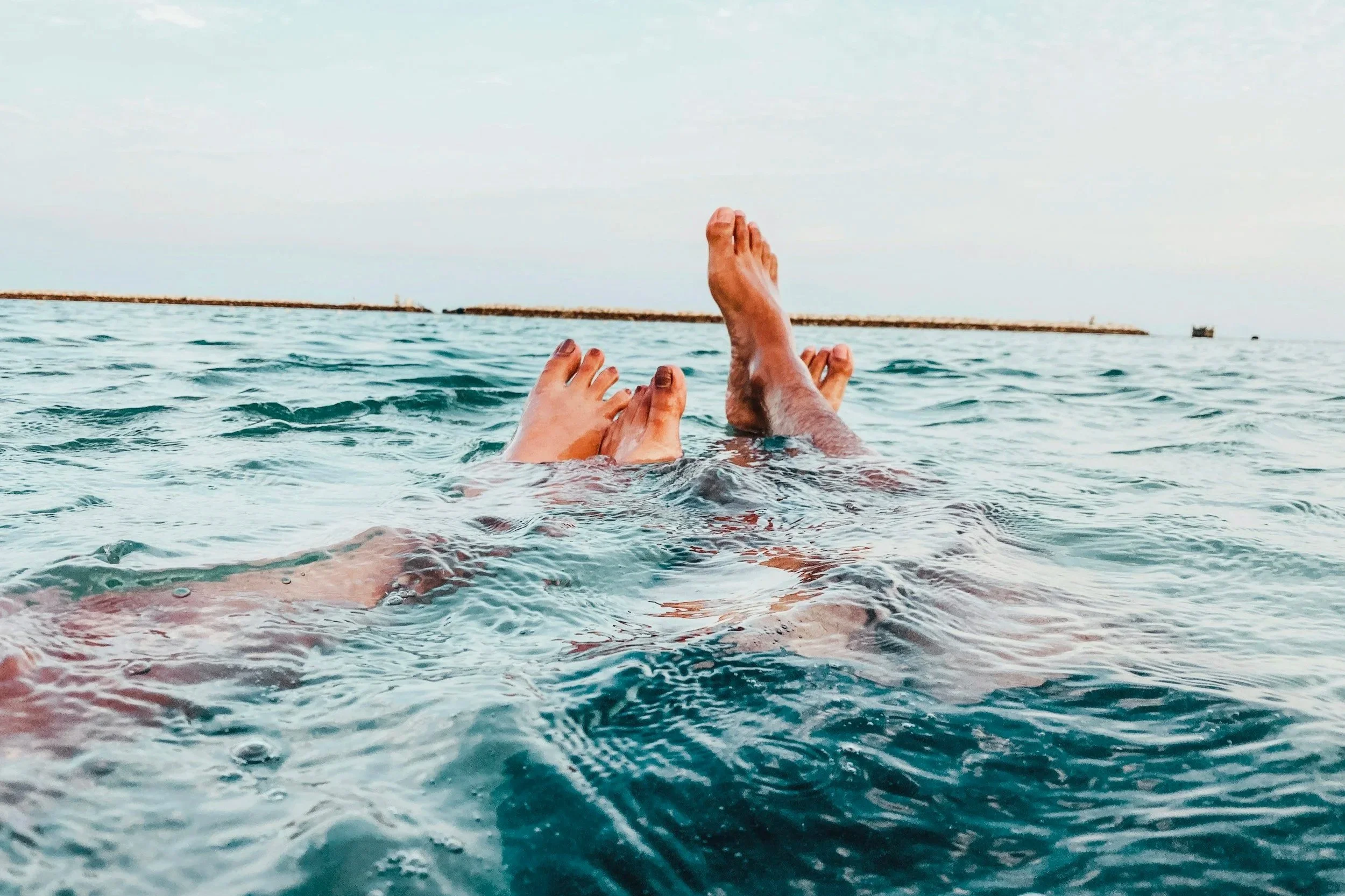 Two pairs of legs and feet are floating in the water at the beach, with a breakwater and boats visible in the distance under a cloudy sky.