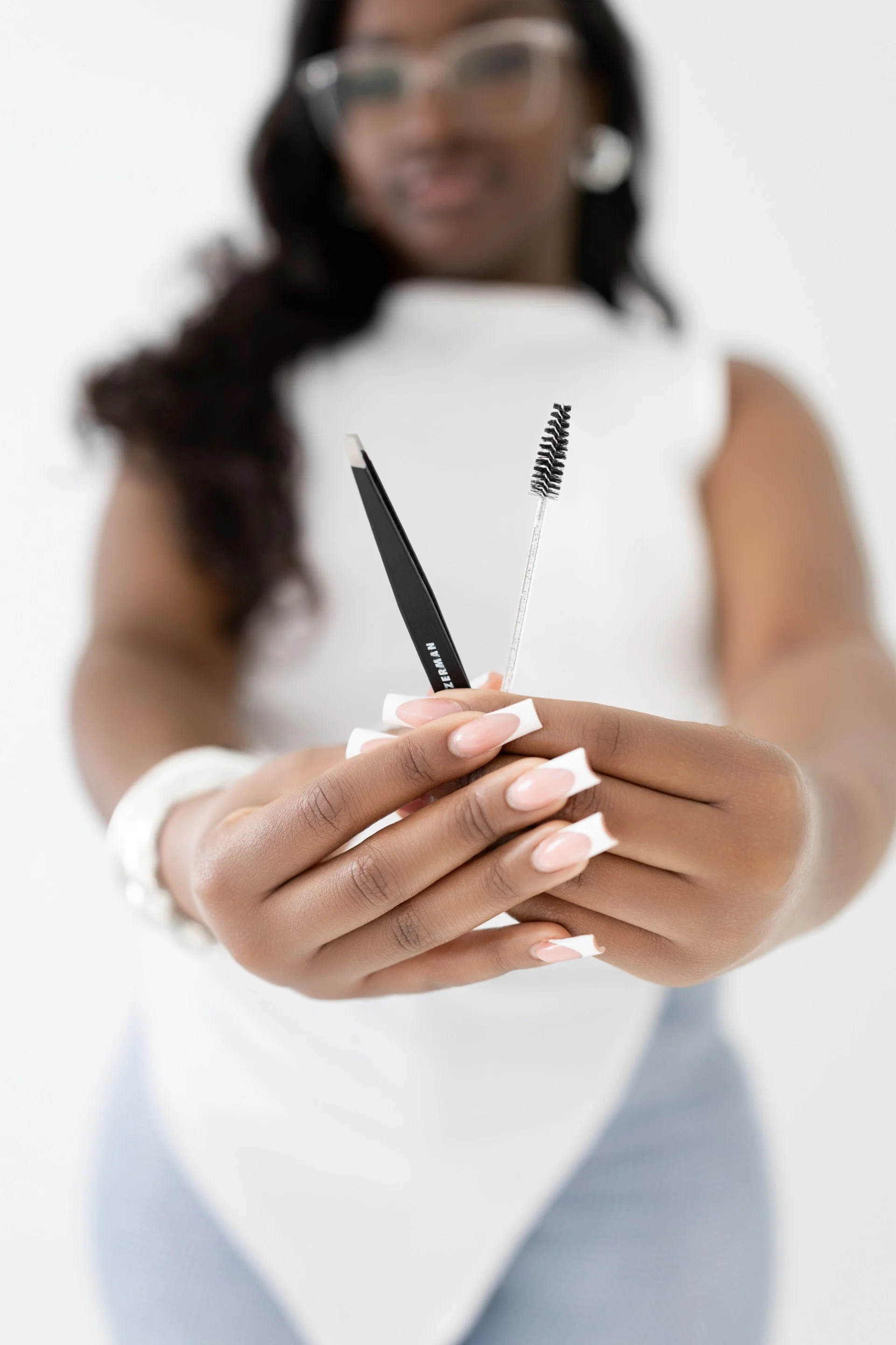 A woman with manicured nails holding eyelash extension tools, including a spoolie brush and a tweezer, close to the camera.
