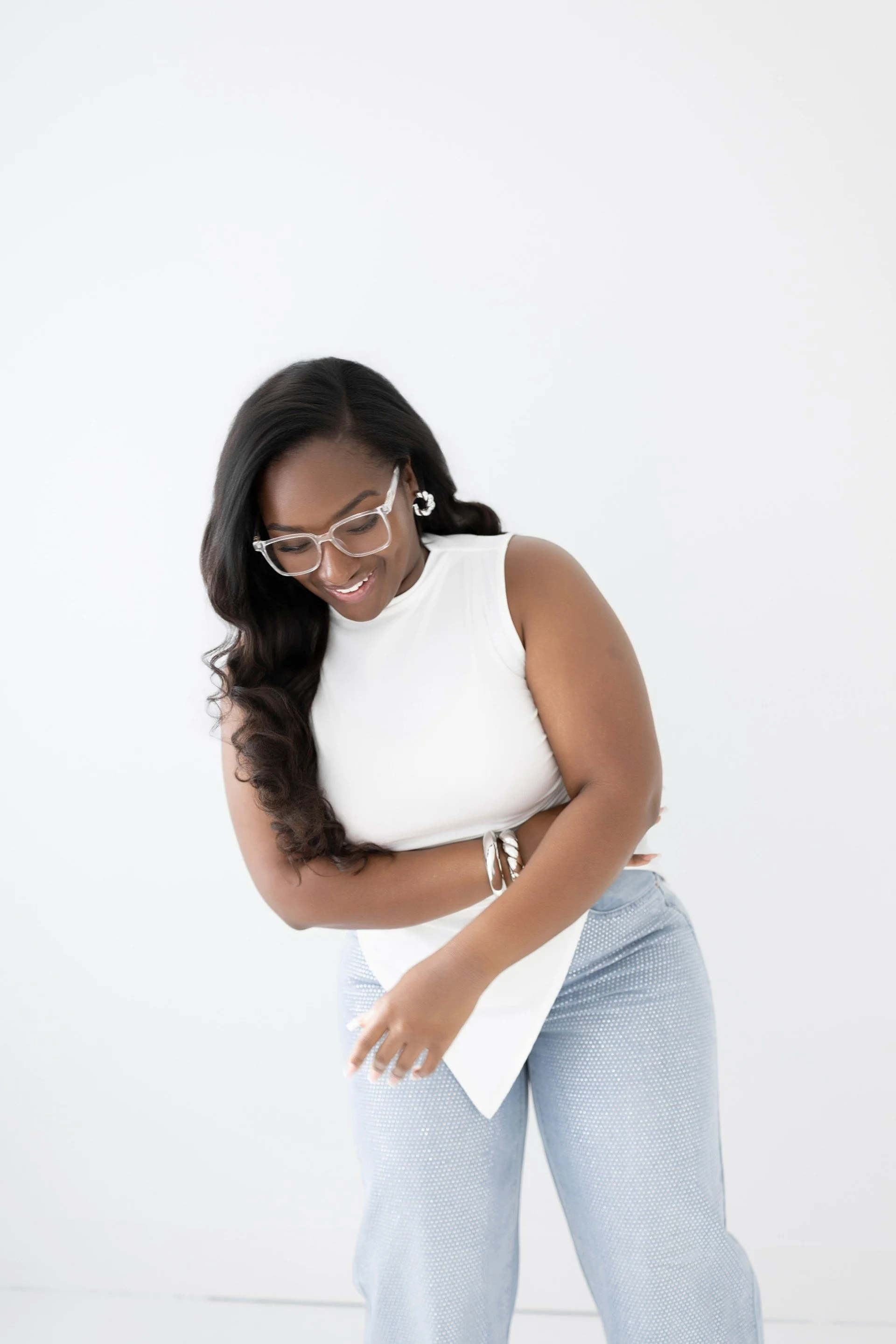 Smiling woman with glasses and long dark hair wearing a white sleeveless top and light blue pants, looking down and laughing.