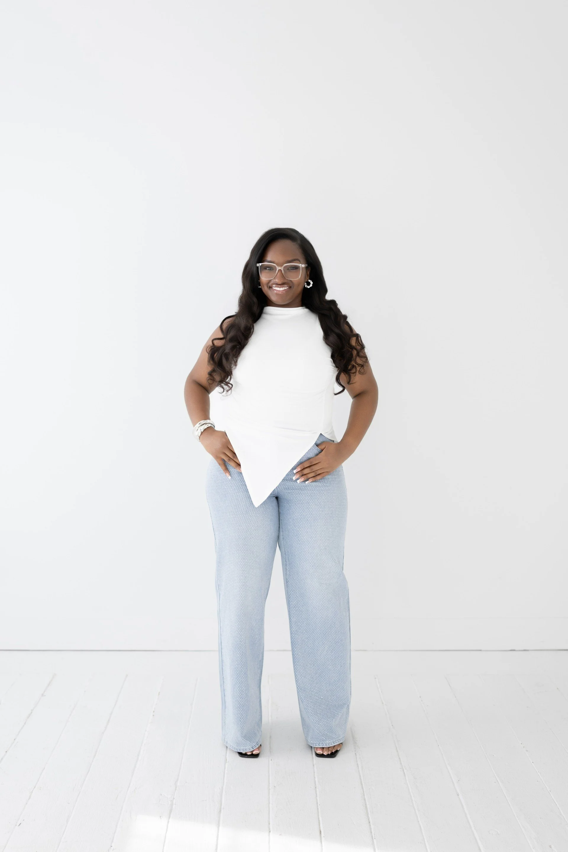 A woman with long dark hair, glasses, and earrings stands with hands on hips in a white sleeveless top and light blue wide-leg pants against a white wall.