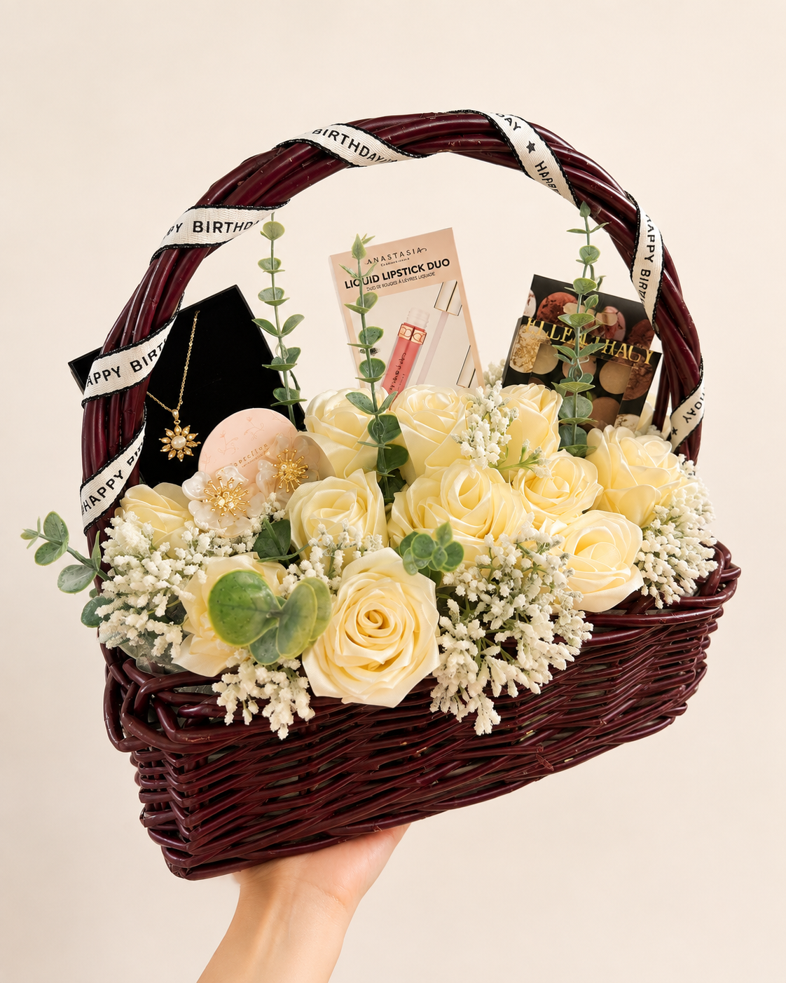 A hand holding a gift basket containing white roses, greenery, and celebratory items with a 'Happy Birthday' ribbon.