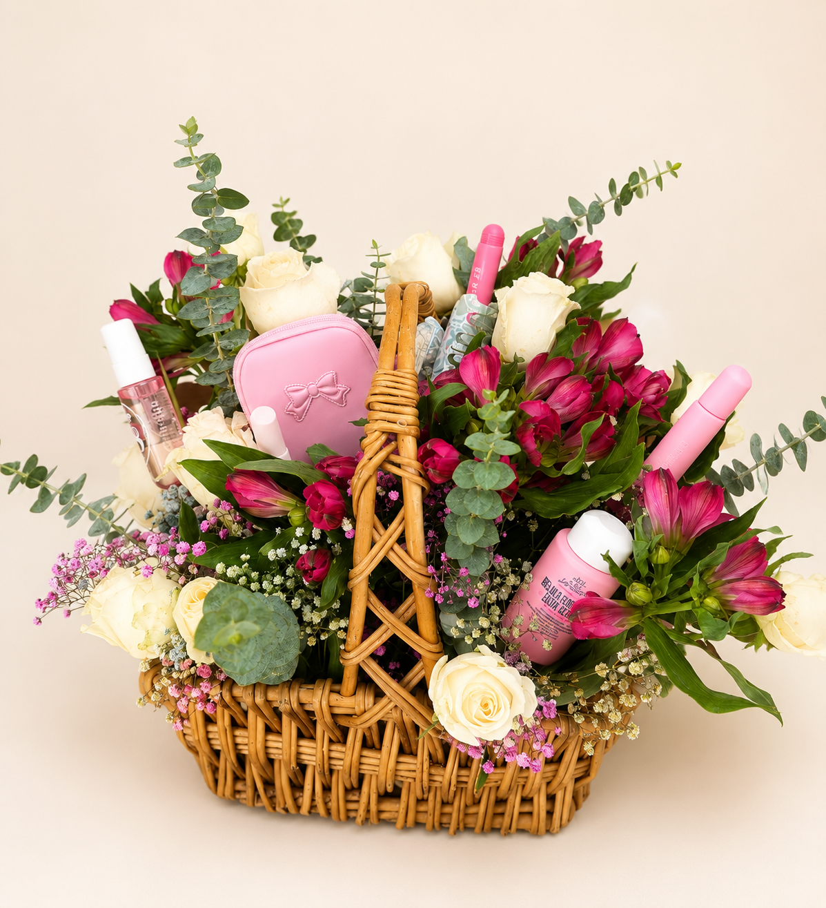 A wicker basket filled with white roses, pink flowers, eucalyptus leaves, and other small flowers, containing personal care items such as pink and white bottles of lotions or sprays, a pink cosmetic pouch, and other toiletries.