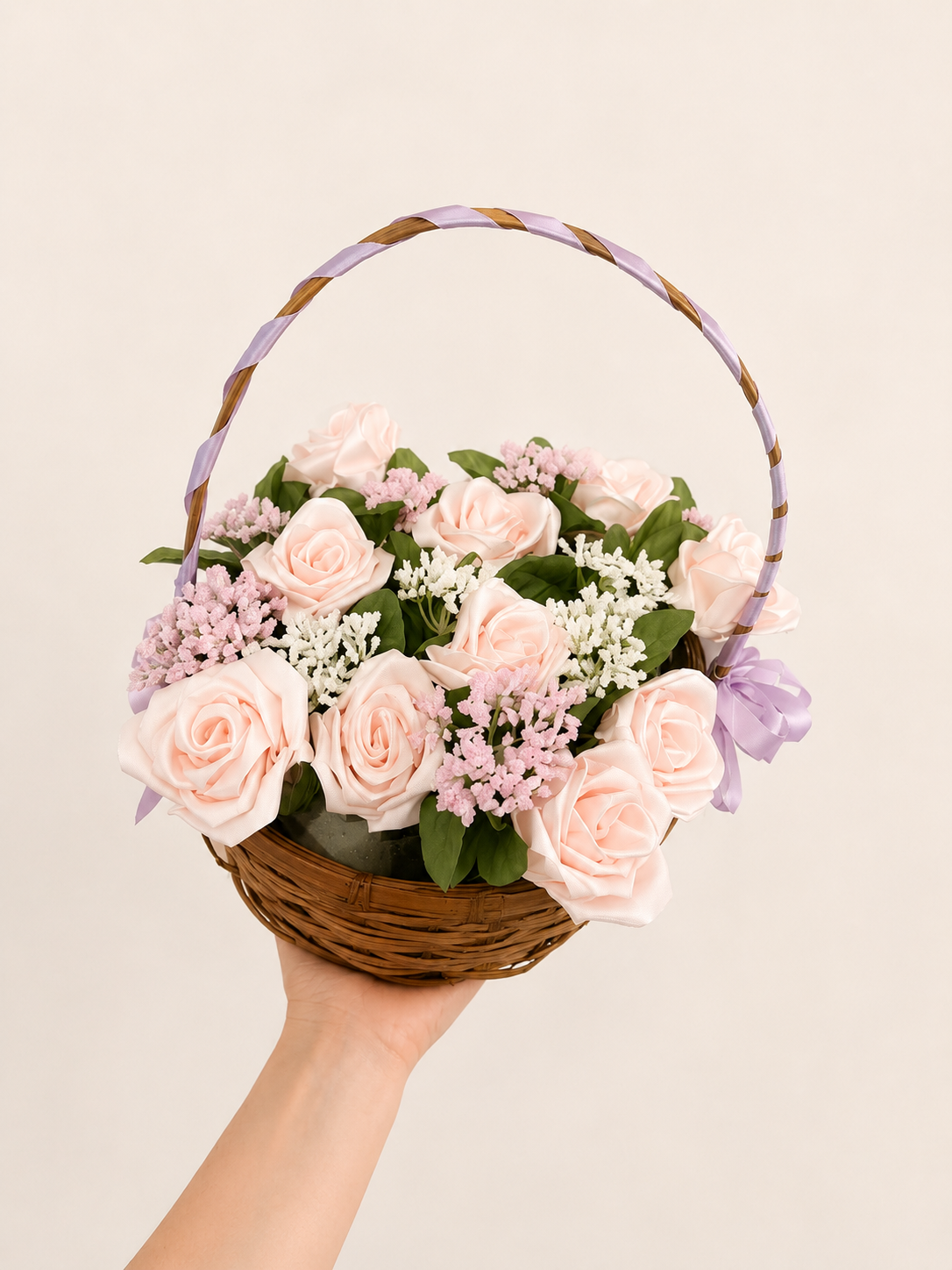 Hand holding a wicker basket filled with pink roses, white and pink filler flowers, and green leaves, decorated with a purple ribbon.