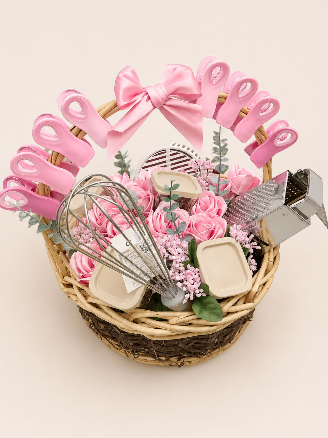 A gift basket filled with pink artificial roses, kitchen utensils, a cheese grater, a whisk, small containers, eucalyptus, pink flowers, and a pink bow on the handle of a pink silicone spatula.