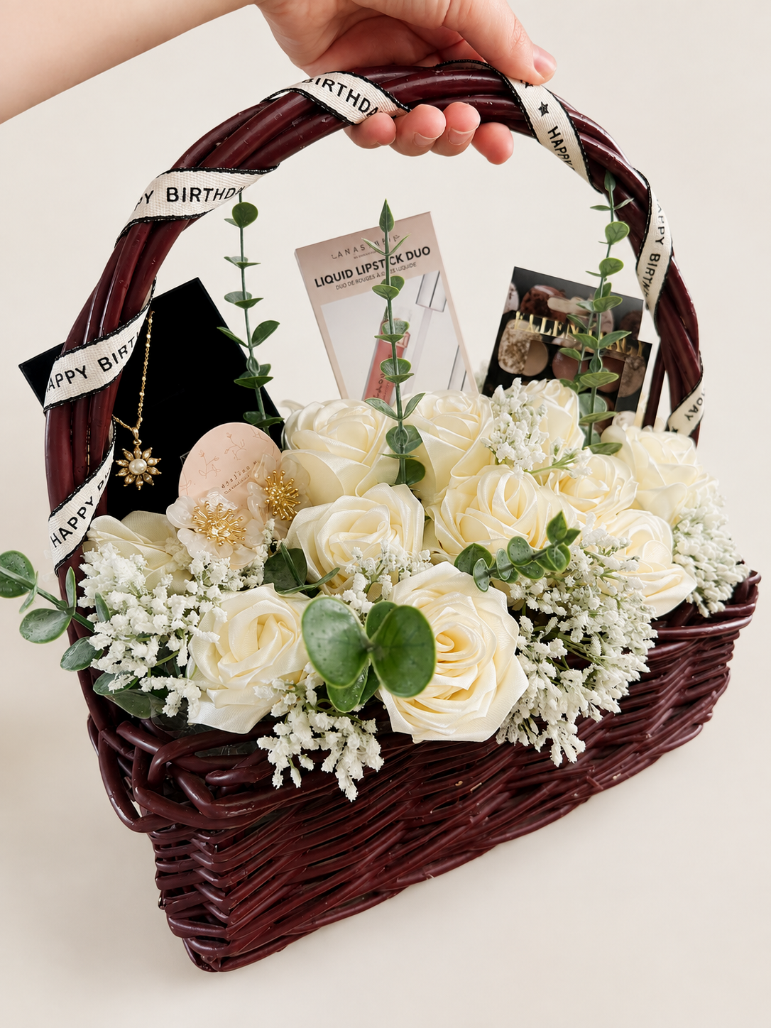 A hand holding a burgundy woven basket decorated with a "Happy Birthday" ribbon, filled with white roses, greenery, and small white flowers, along with skincare and makeup products and jewelry.