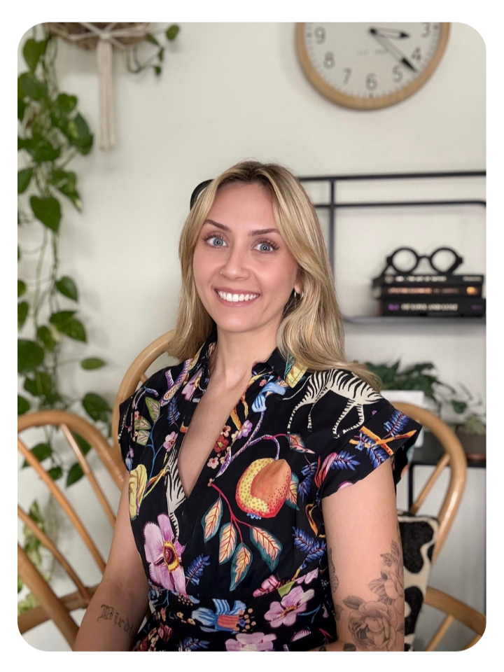 A woman with blonde hair smiling at the camera, wearing a black floral dress, sitting in a room with a clock and bookshelf in the background, and plants around her.