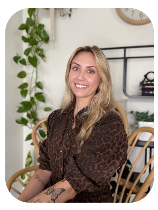 A woman with long blonde hair, wearing a brown leopard print shirt, sitting indoors with a smile. In the background, there are green houseplants, a clock, and a black shelf with books and decorative items.