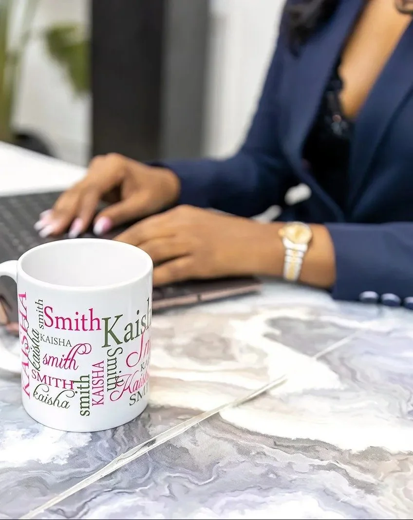 A white coffee mug with various names including 'Kaisi', 'Smith', and 'Kaisha' printed in different fonts and colors on a marble table. A woman wearing a dark blue blazer and gold watch is working on a laptop in the background.