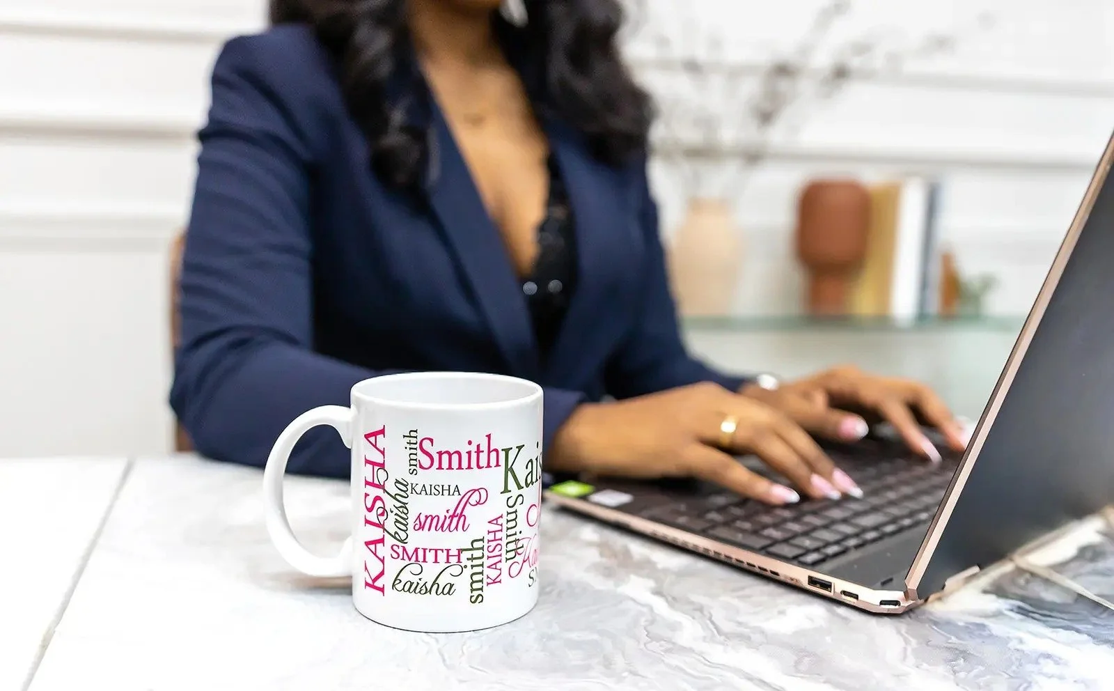 A woman dressed in a dark blazer working on a laptop at a marble table with a white mug featuring colorful names.