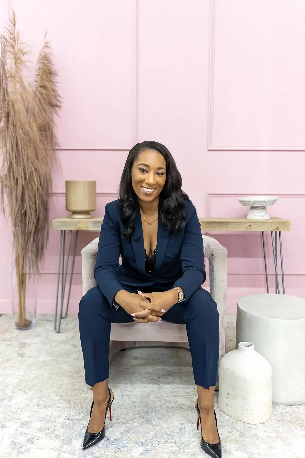 A woman in a navy blue suit sitting on a beige armchair in a modern, pink-walled room. She has black wavy hair, wearing heels, and smiling at the camera. There are decorative objects and a tall pampas grass plant behind her.