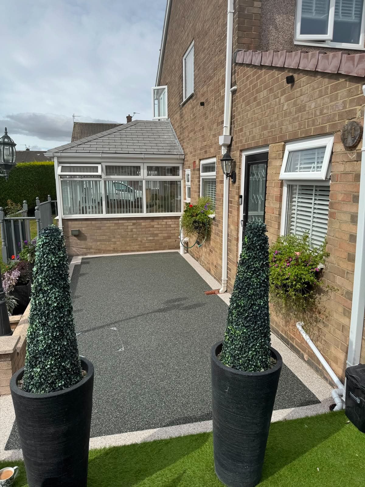 A small backyard patio with two tall potted plants, artificial grass, and a brick house with a window and an outdoor lantern.