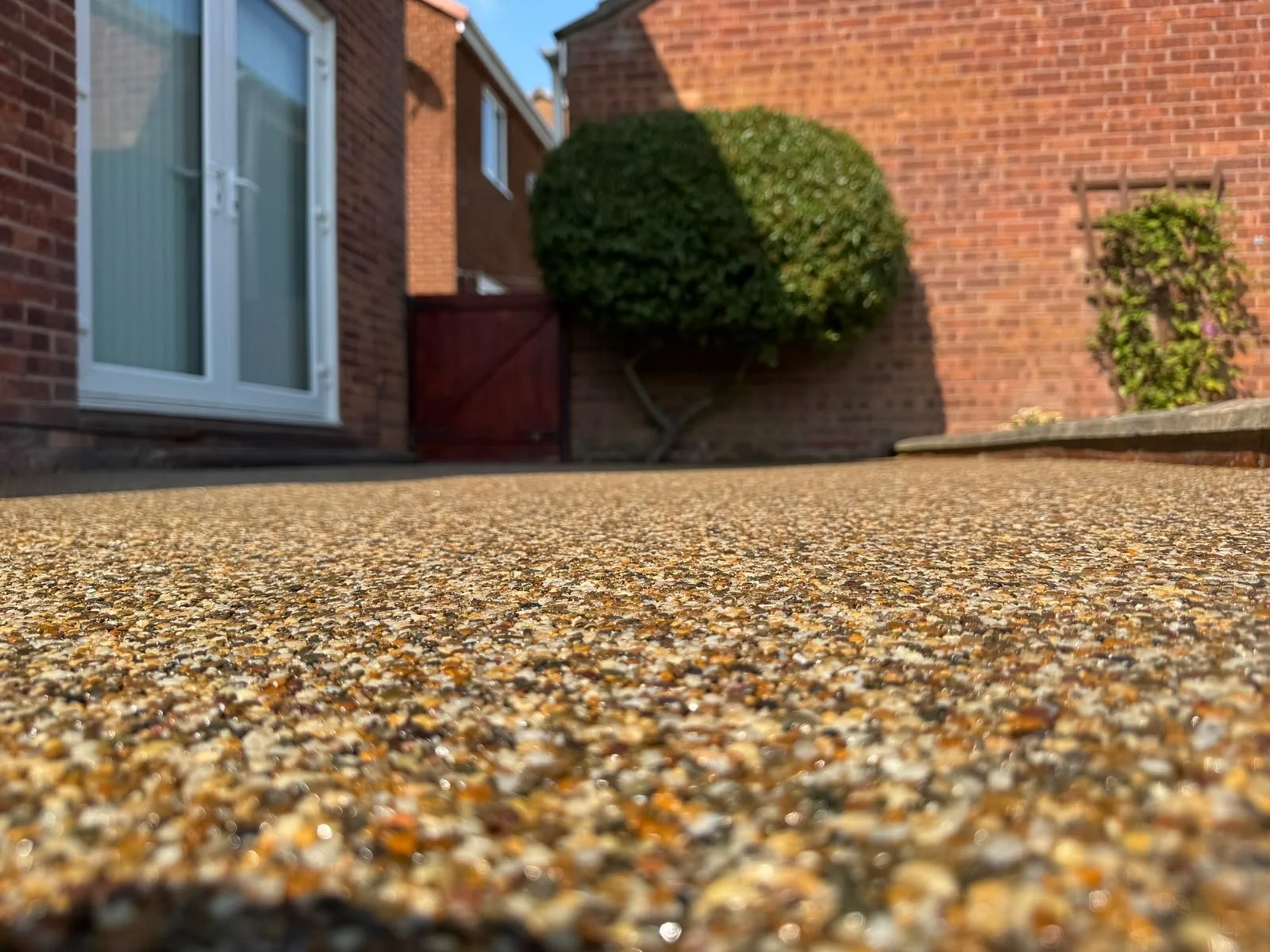 Close-up of a textured pebble driveway with brick houses, green bushes, and a red wooden fence in the background.