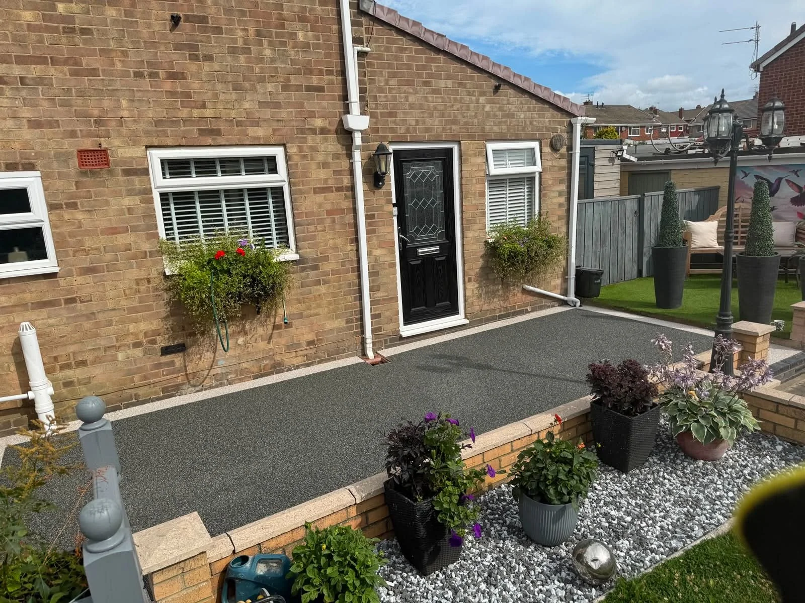 A backyard with a brick house, two windows with white blinds, black door with decorative glass, and various potted plants on a gravel and paved surface.