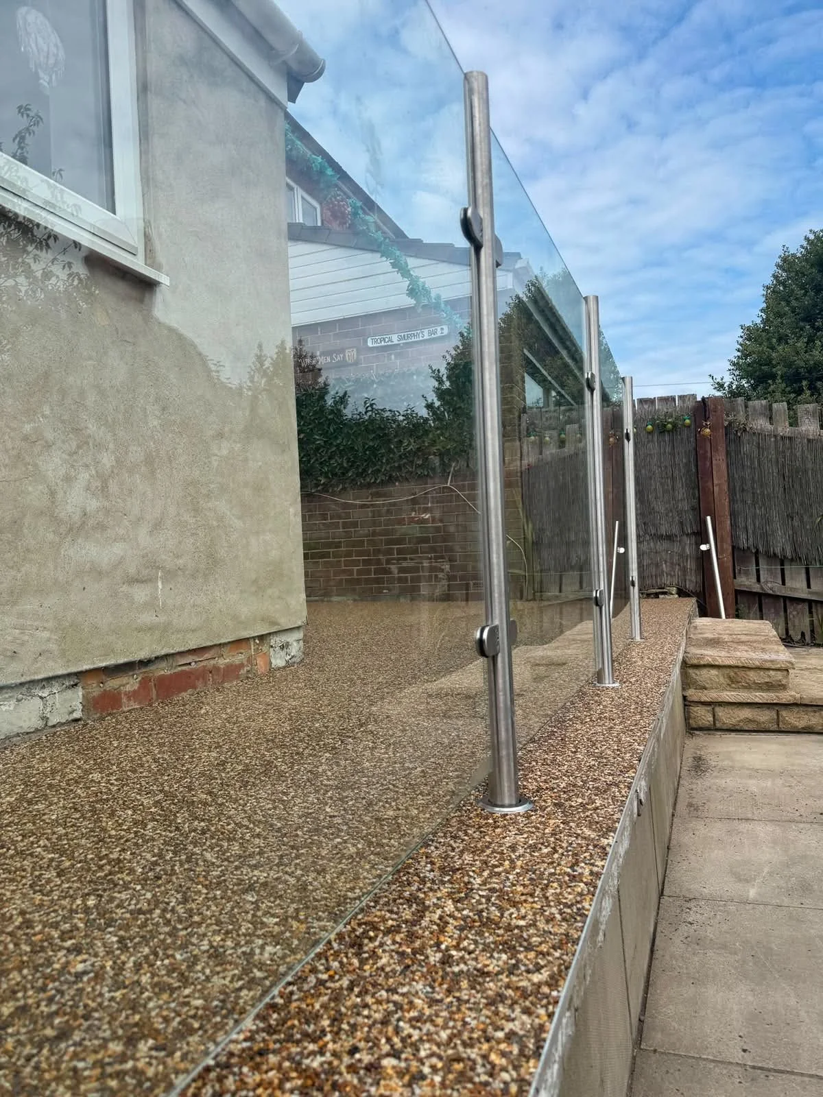 A newly installed glass railing with metal posts on a textured concrete patio outside a house, with a partial view of the backyard and sky in the background.