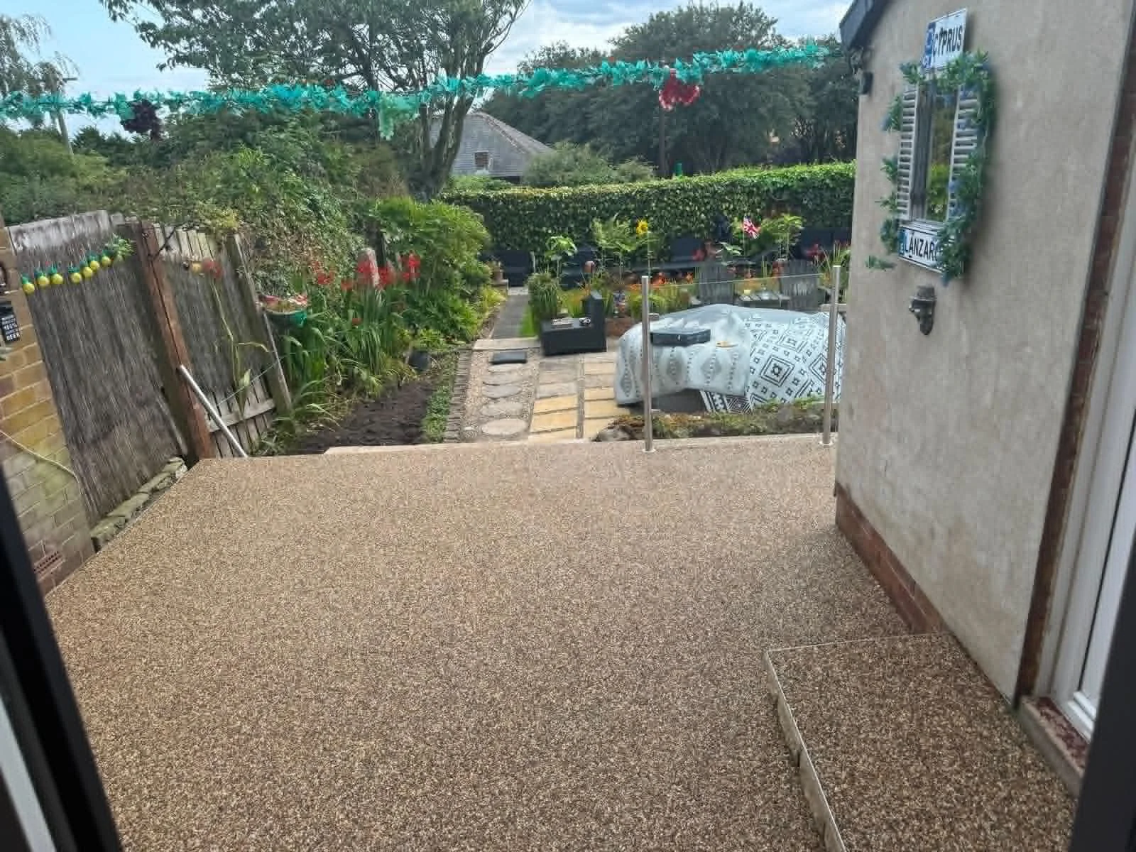 View from inside a house looking out to a garden with a stone pathway, plants, and outdoor furniture, decorated with holiday ornaments and a string of greenery.