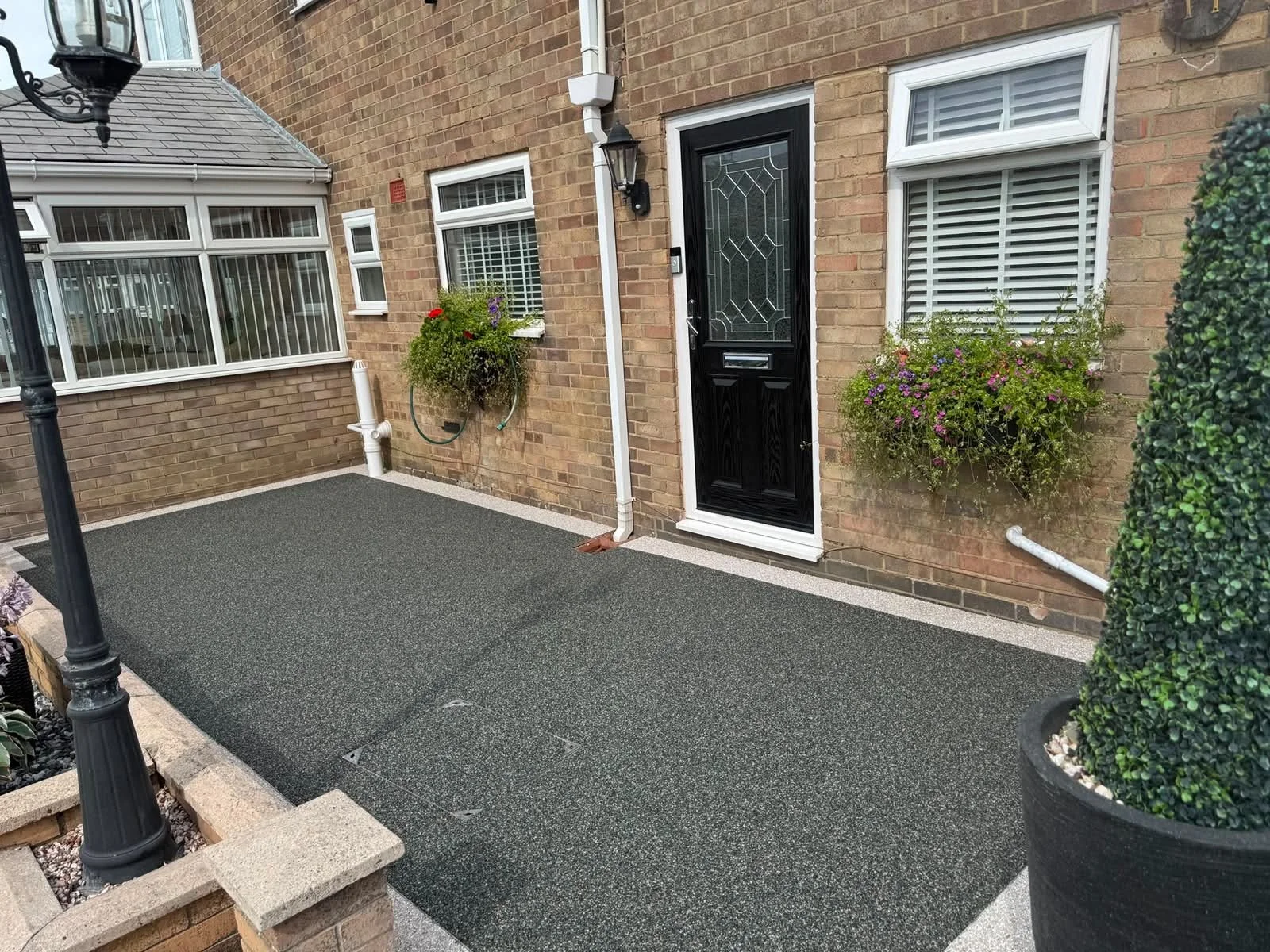 A small outdoor porch area with a black front door, two windows with white blinds, potted plants with flowers, and a black lamp post on the left.