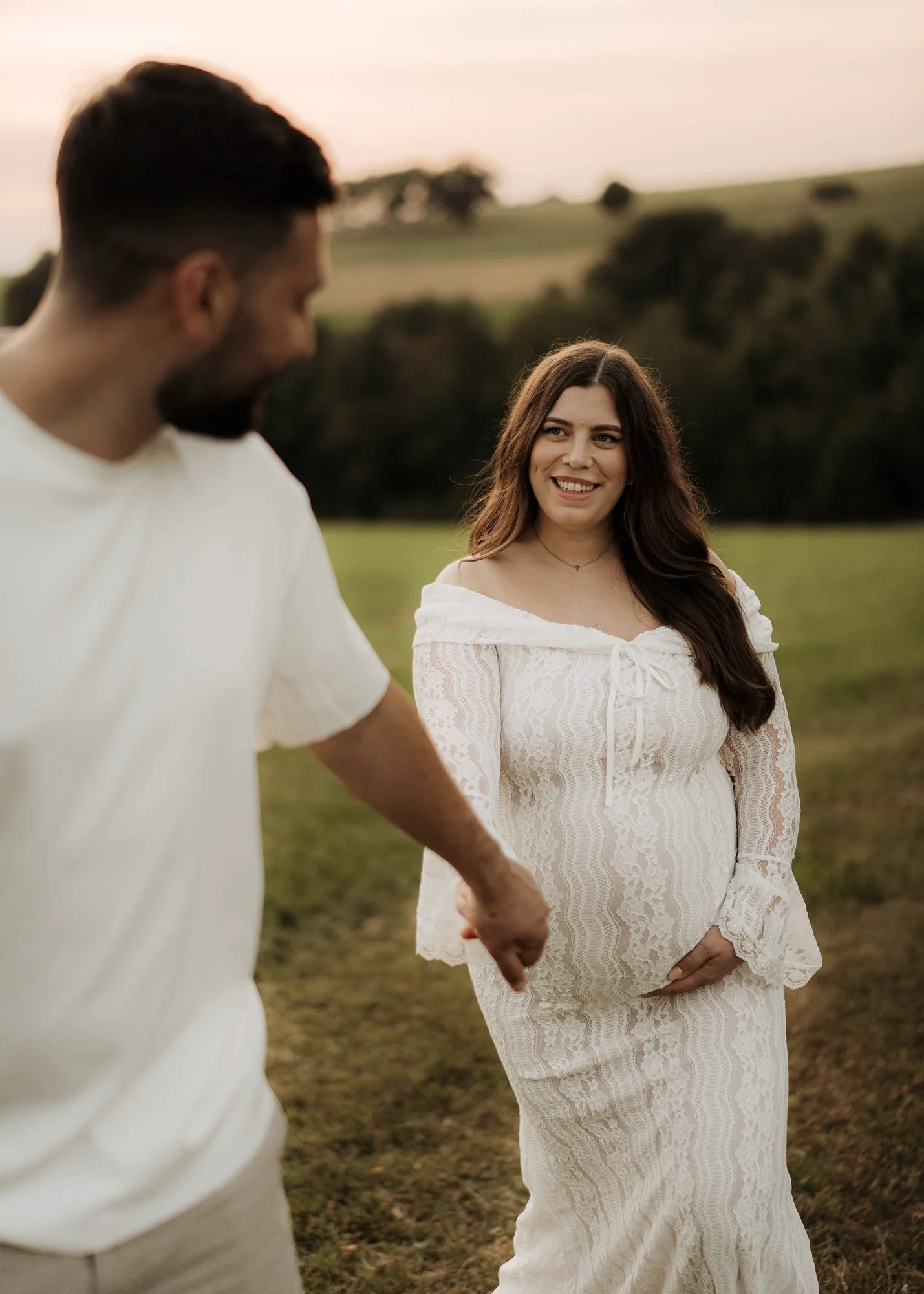 Ein Paar, das gemeinsam im Freien spaziert. Die Frau trägt ein weißes Spitzenkleid und schaut lächelnd nach vorne, während der Mann in einem weißen Shirt sie mit sich nach vorne zieht und zu ihr nach hinten schaut.