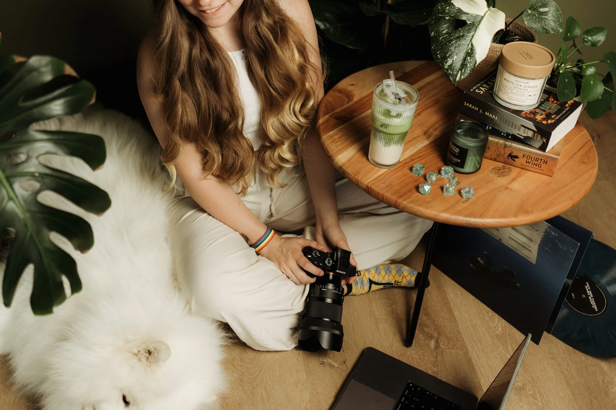 Eine junge Frau mit langen Haaren sitzt auf dem Boden vor einem Tisch, hält eine Kamera in der Hand, neben ihr ein weißer, flauschiger Hund. Auf dem Tisch liegen Bücher, eine Kerze, ein Matchaglas, einige Würfel und eine Vinylplatte.