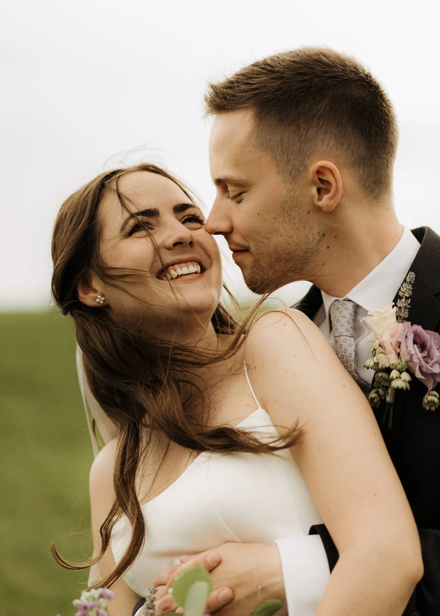 Ein locker lachendes Paar bei einer Hochzeit, wobei der Mann die Frau von hinten umarmt und sie in einem offenen Feld mit hellem Himmel stehen.