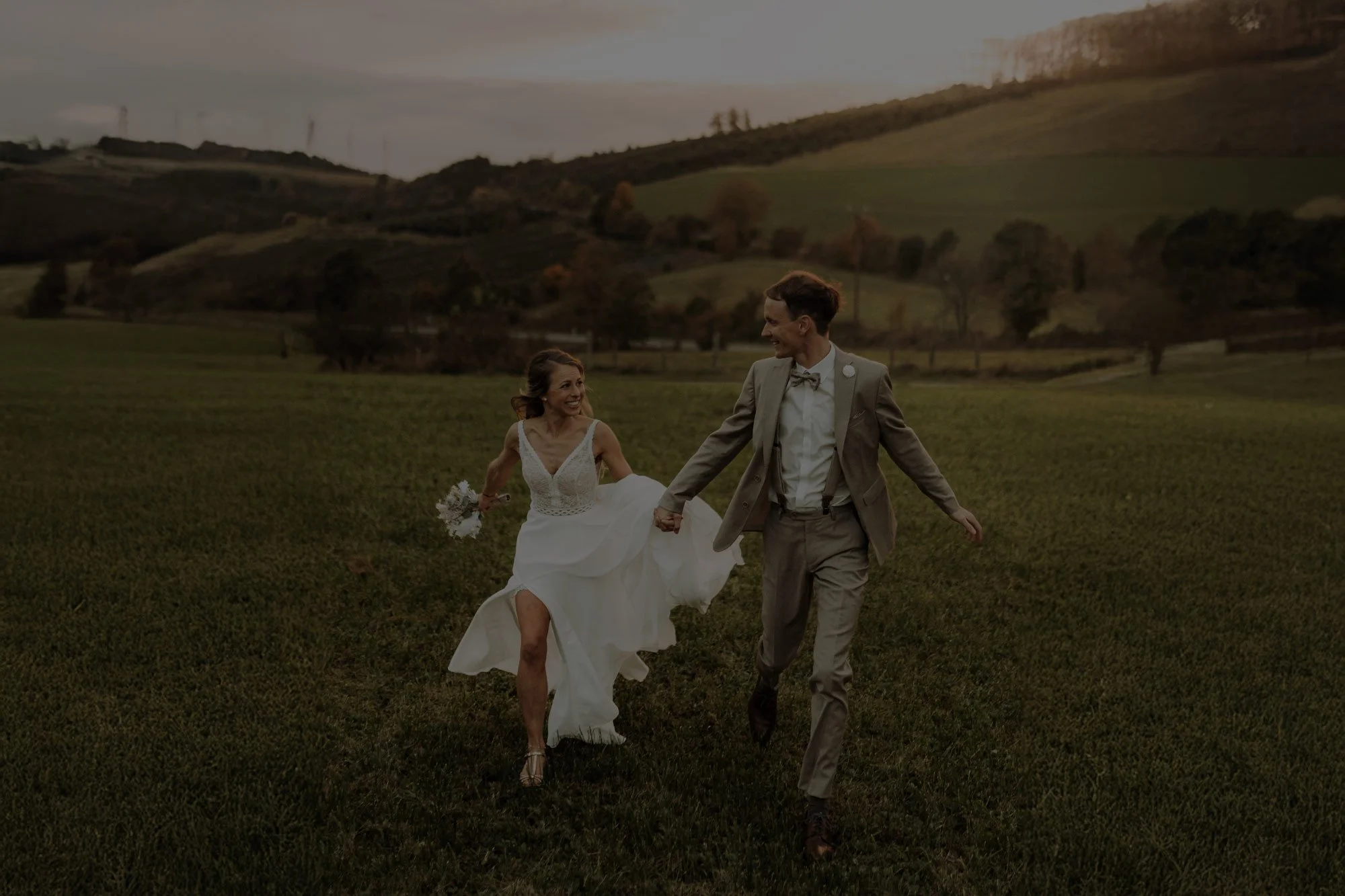 Ein Brautpaar läuft lachend und sich anschauend über eine grüne Wiese bei Sonnenuntergang.