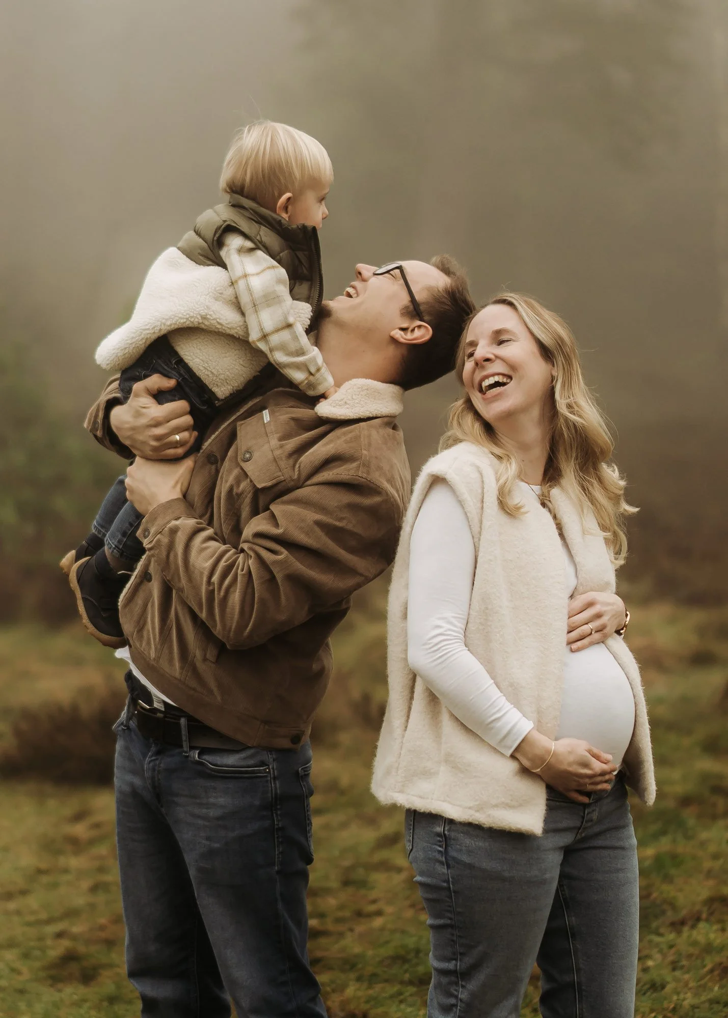 Ein glückliches Paar mit einem Kind im Freien in einem Wald, die Frau ist schwanger, der Mann hebt das Kind hoch, alle lachen und genießen die gemeinsame Zeit.