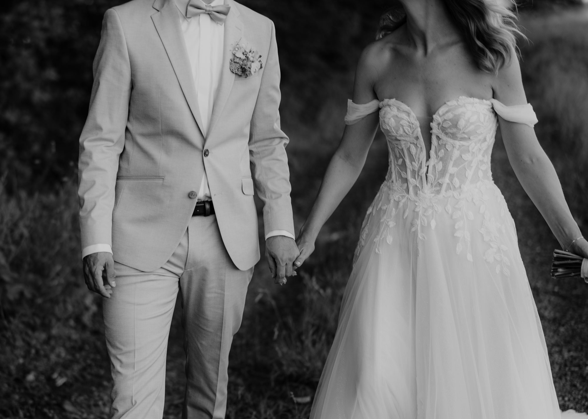 Schwarz-weiß-Foto eines Paares bei ihrer Hochzeit, in der sie ihre Hände halten, die Braut trägt ein detailliertes, trägerloses Hochzeitskleid und der Bräutigam einen hellen Anzug mit Fliege.