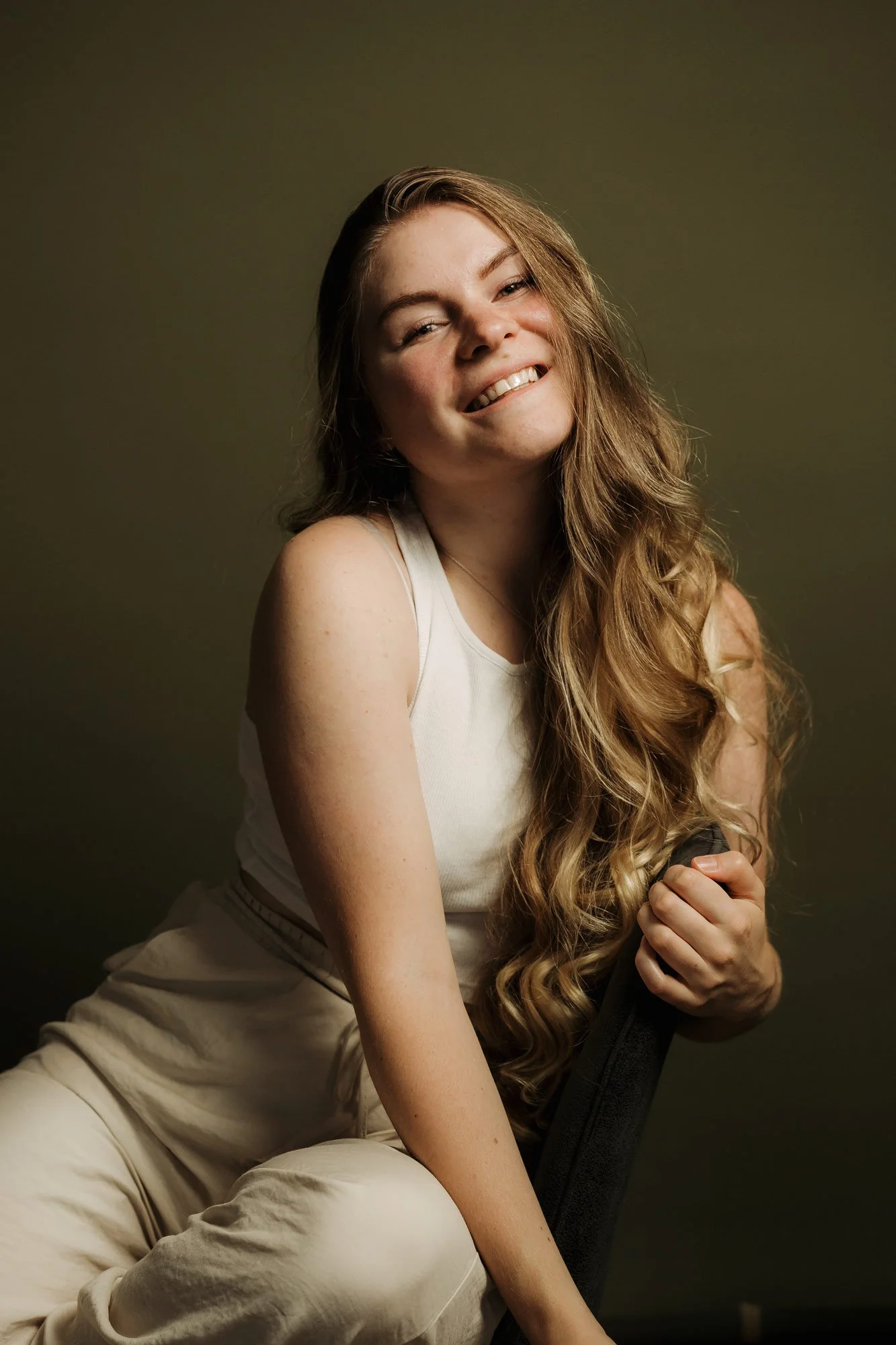 Junge Frau mit langen, lockigen, blonden Haaren, lacht und sitzt locker schräg auf einem Stuhl. Sie trägt ein weißes Tanktop und eine helle Hosen vor einem dunkelgrünen Hintergrund.