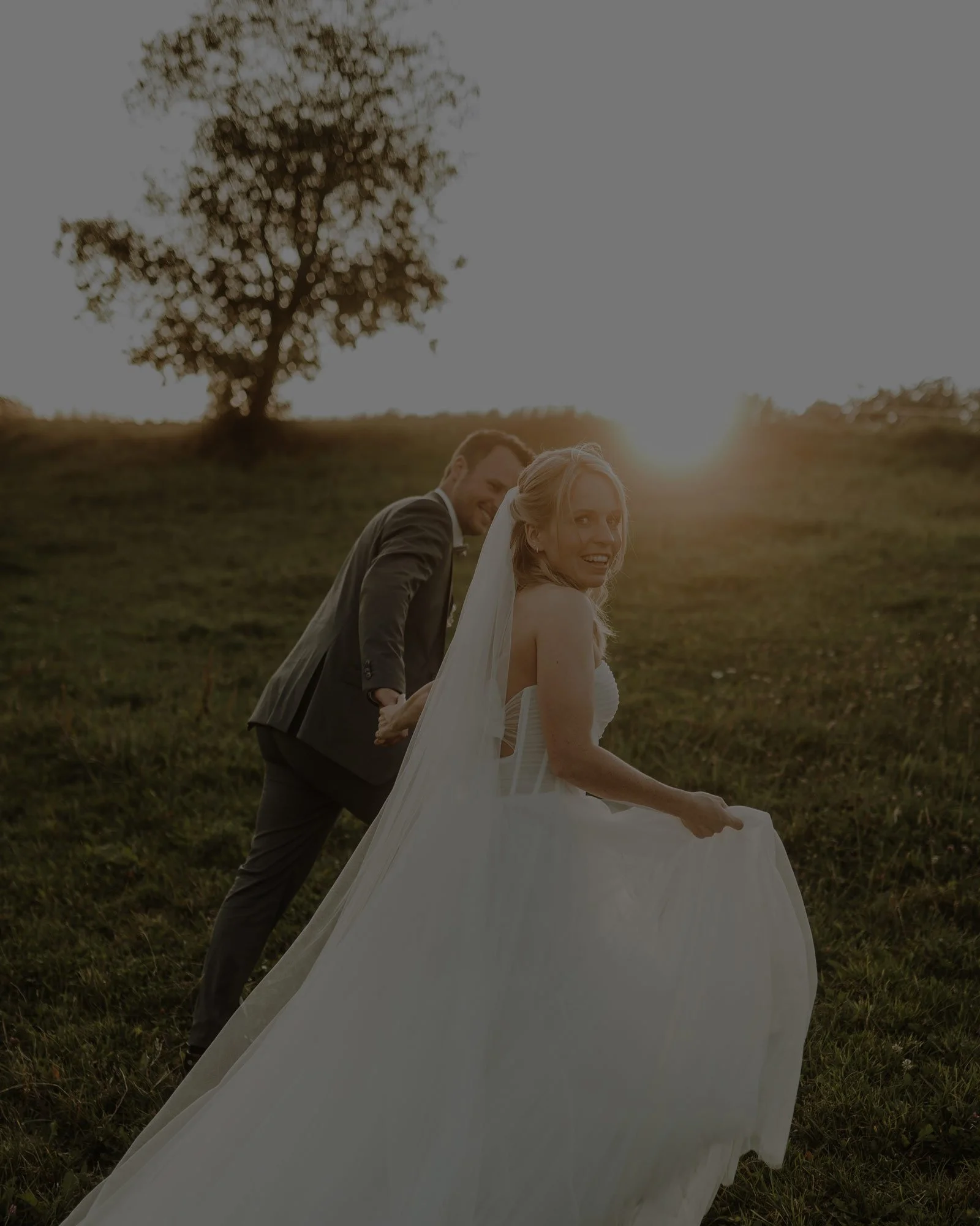 Ein Brautpaar beim Spazieren im Freien bei Sonnenuntergang, die Braut trägt ein langes weißes Kleid und ihr Mann hält ihre Hand. Sie laufen von der Kamera weg, aber die Frau schaut direkt in die Kamera während der Mann sie anschaut.